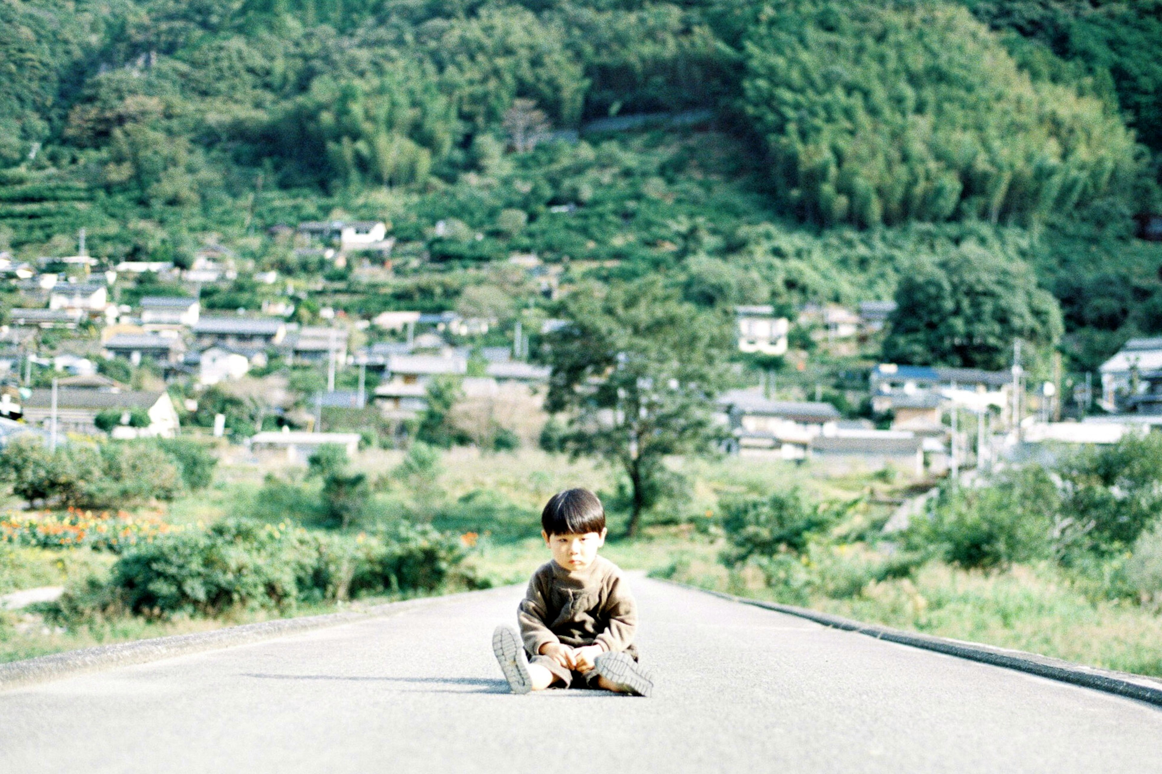 A child sitting on a rural road with green hills and houses in the background