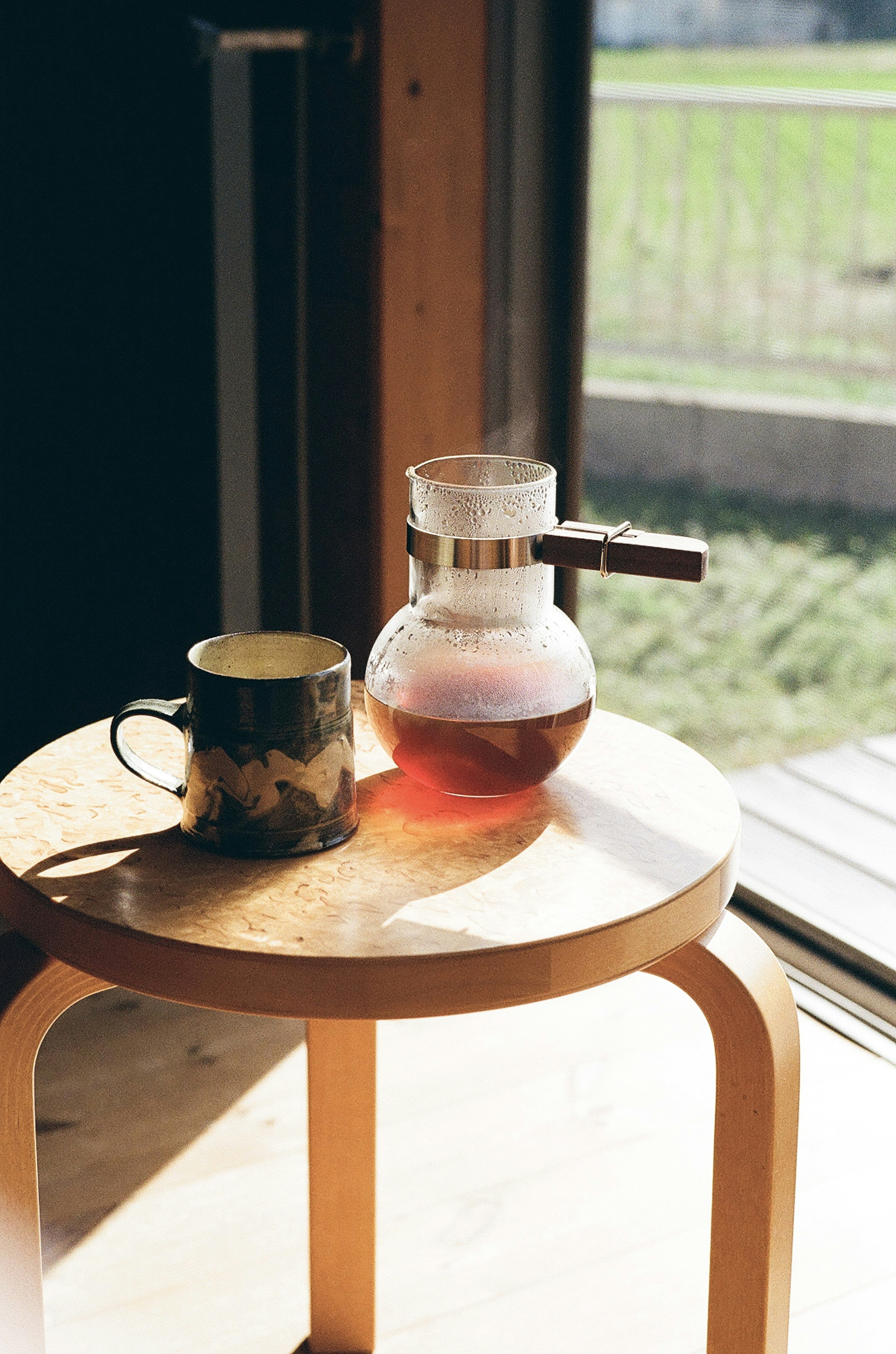 A coffee pot and cup on a wooden stool near a sunlit window