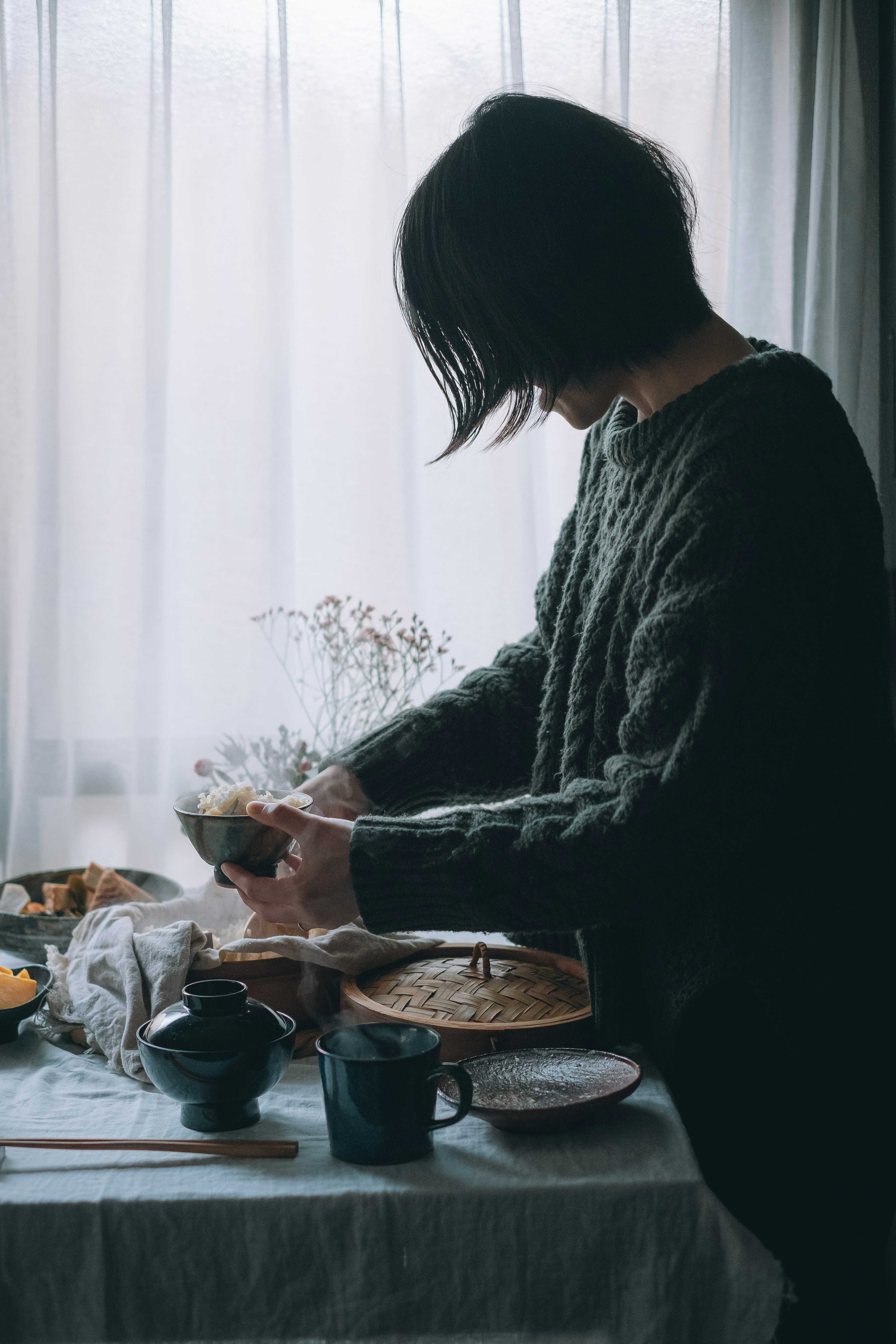 A woman preparing a meal at a table with various dishes and utensils near a window