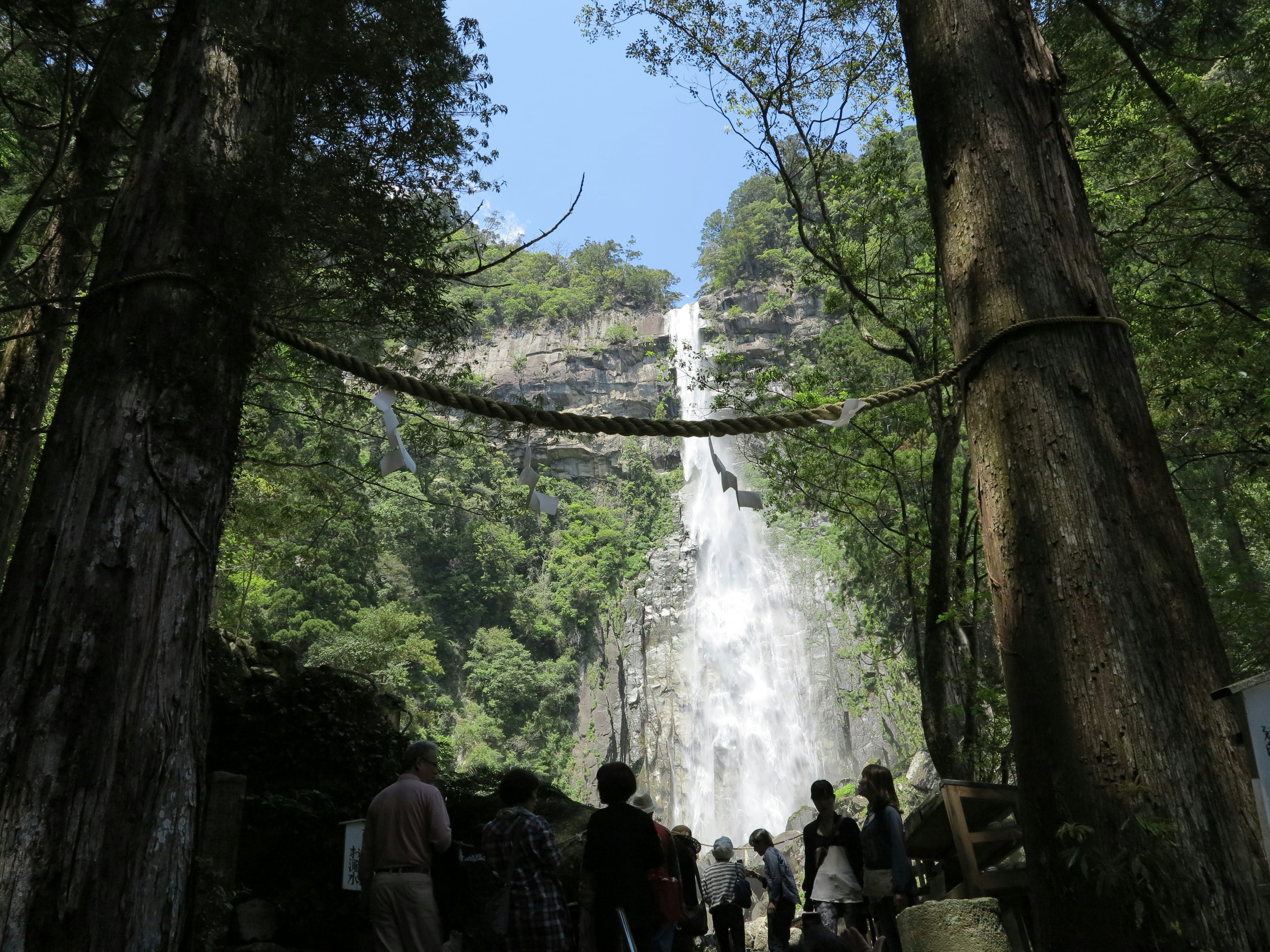 Orang-orang berdiri di depan air terjun dikelilingi pepohonan hijau dan langit biru