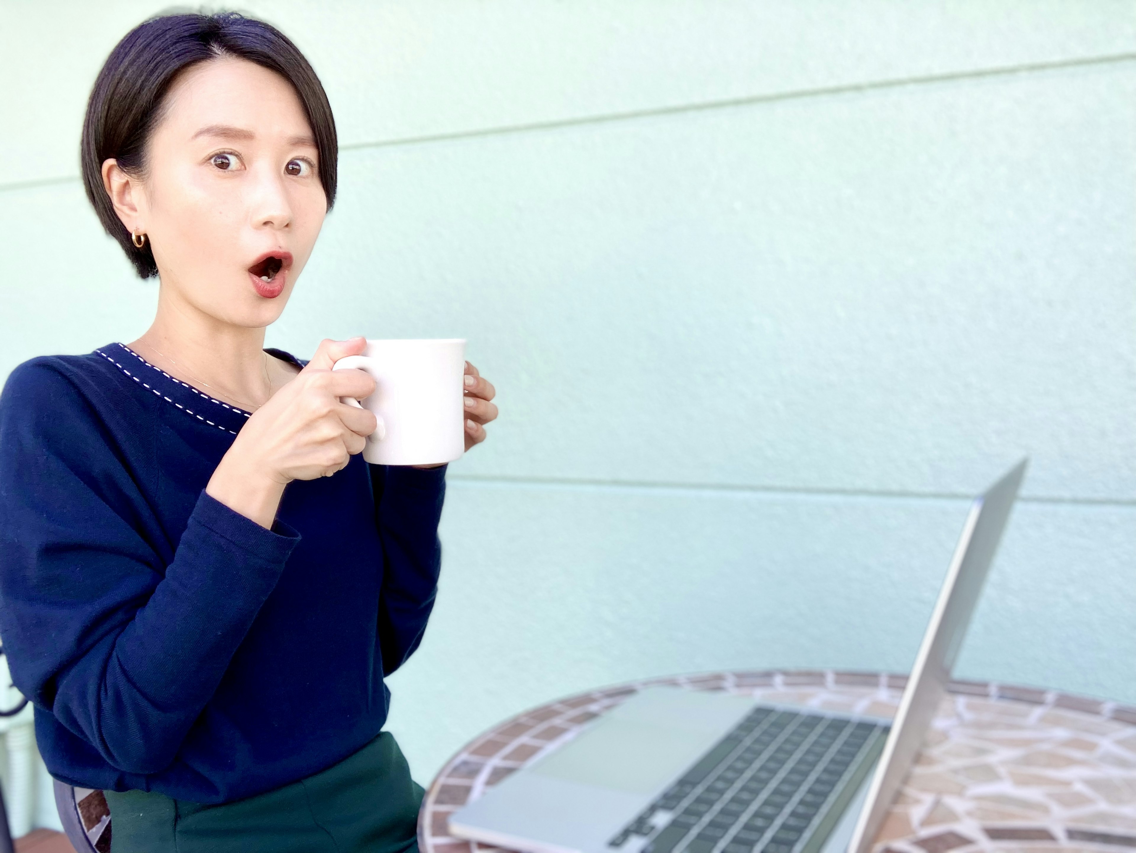 Woman with surprised expression holding a coffee cup sitting in front of a laptop