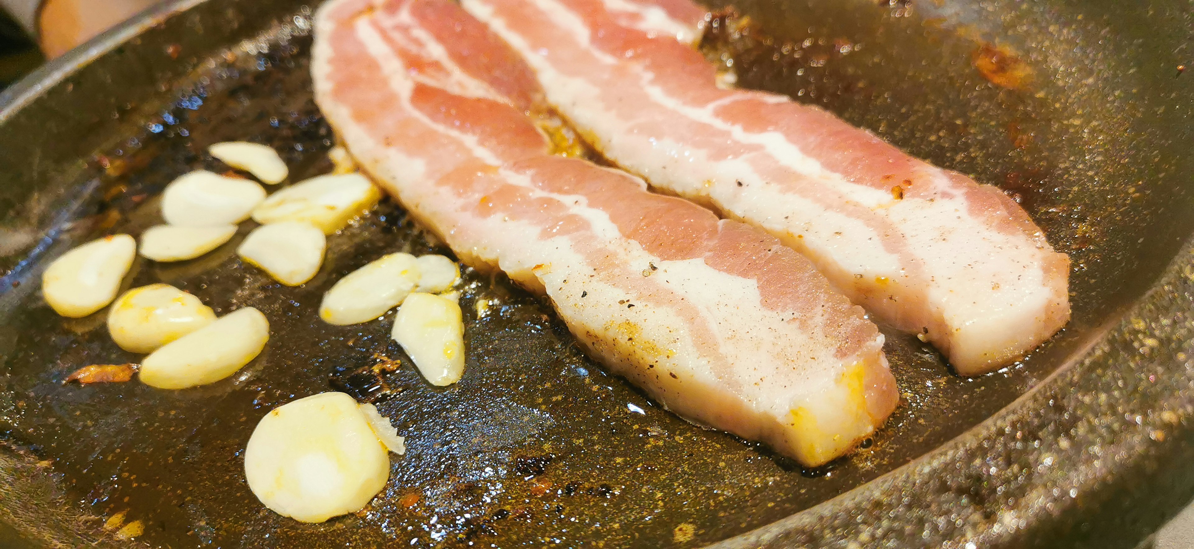 Sizzling bacon strips alongside garlic cloves on a frying pan