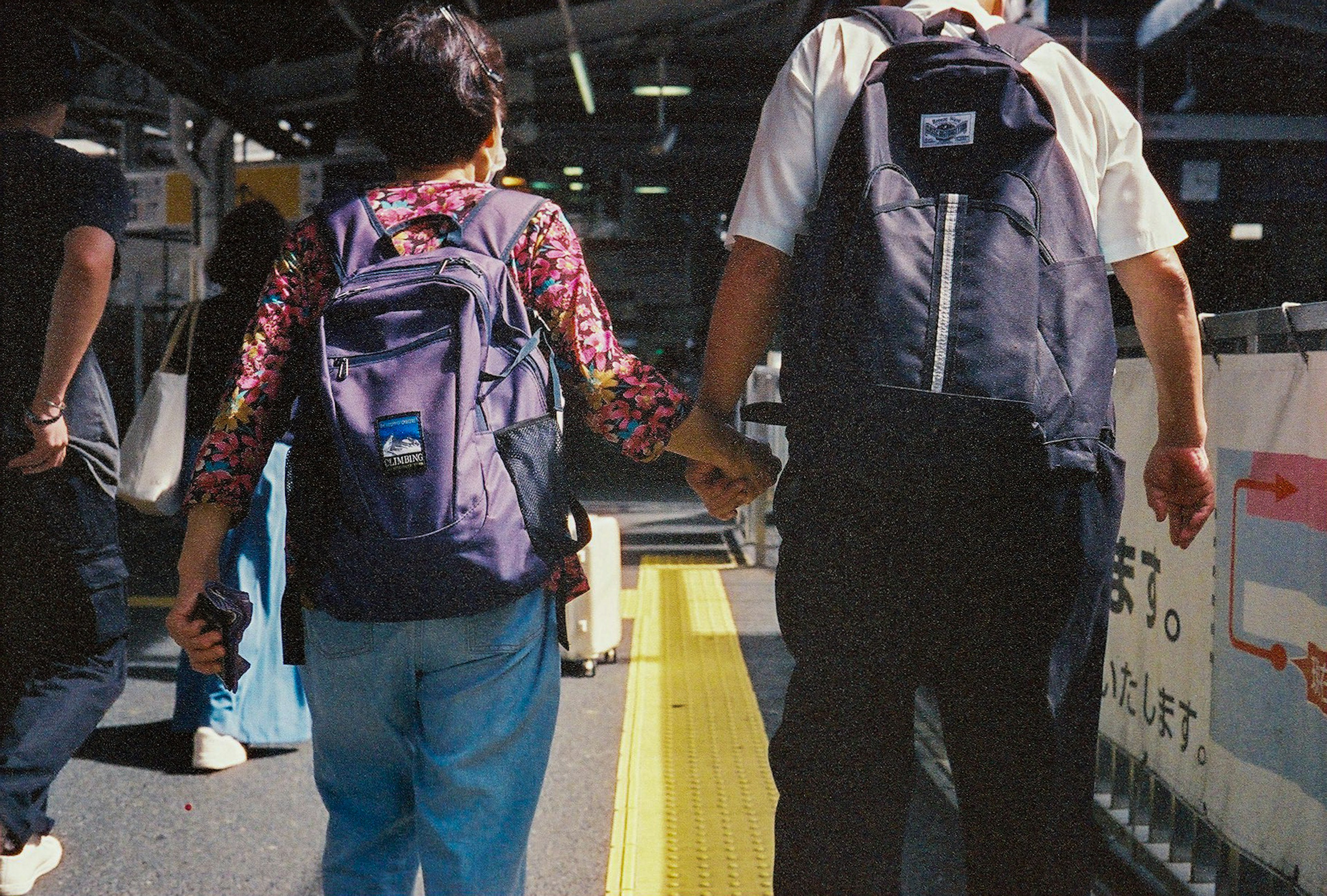 Child and adult holding hands from behind on a train platform wearing purple backpacks