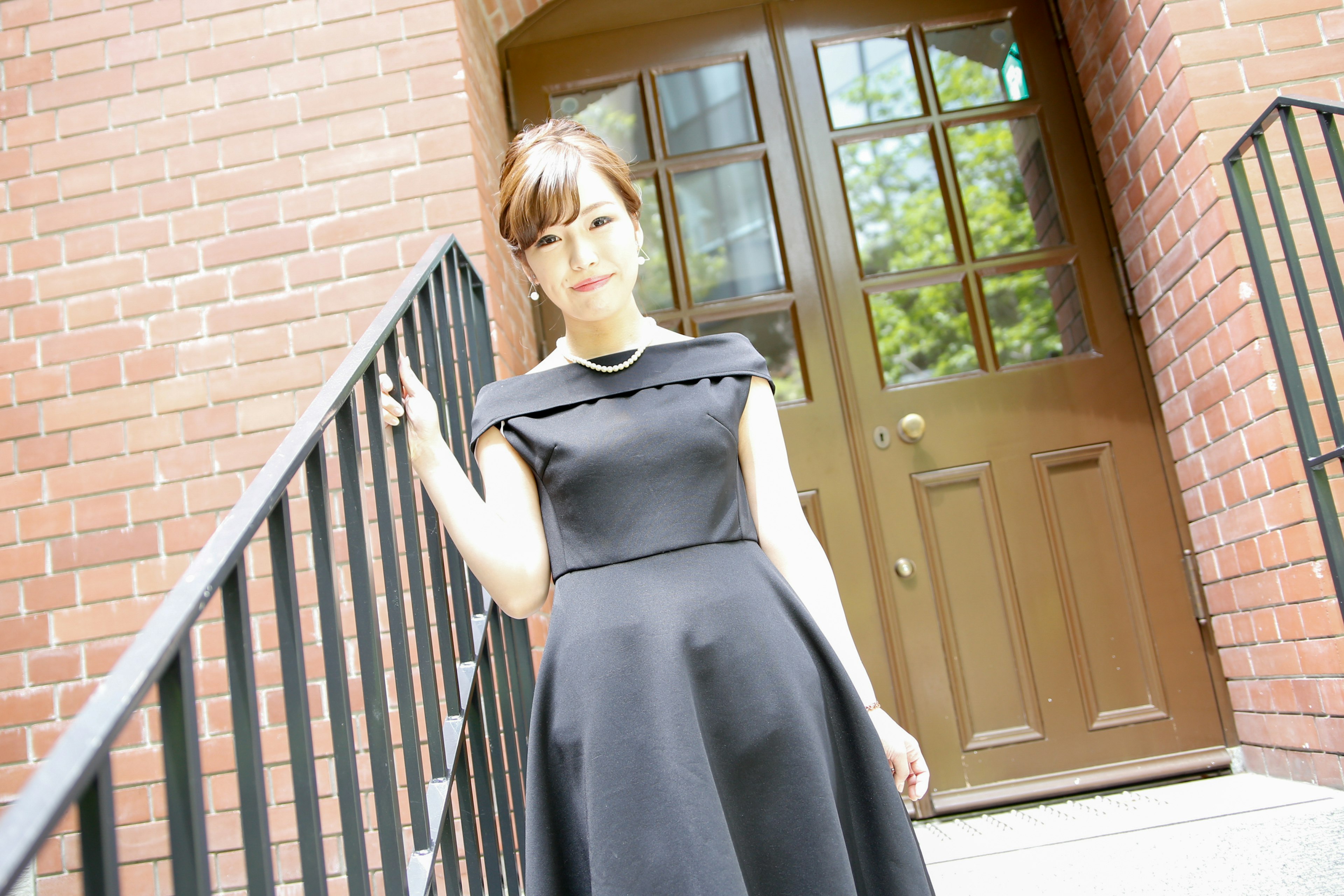 A woman in a black dress stands on stairs with a brick building and wooden door in the background