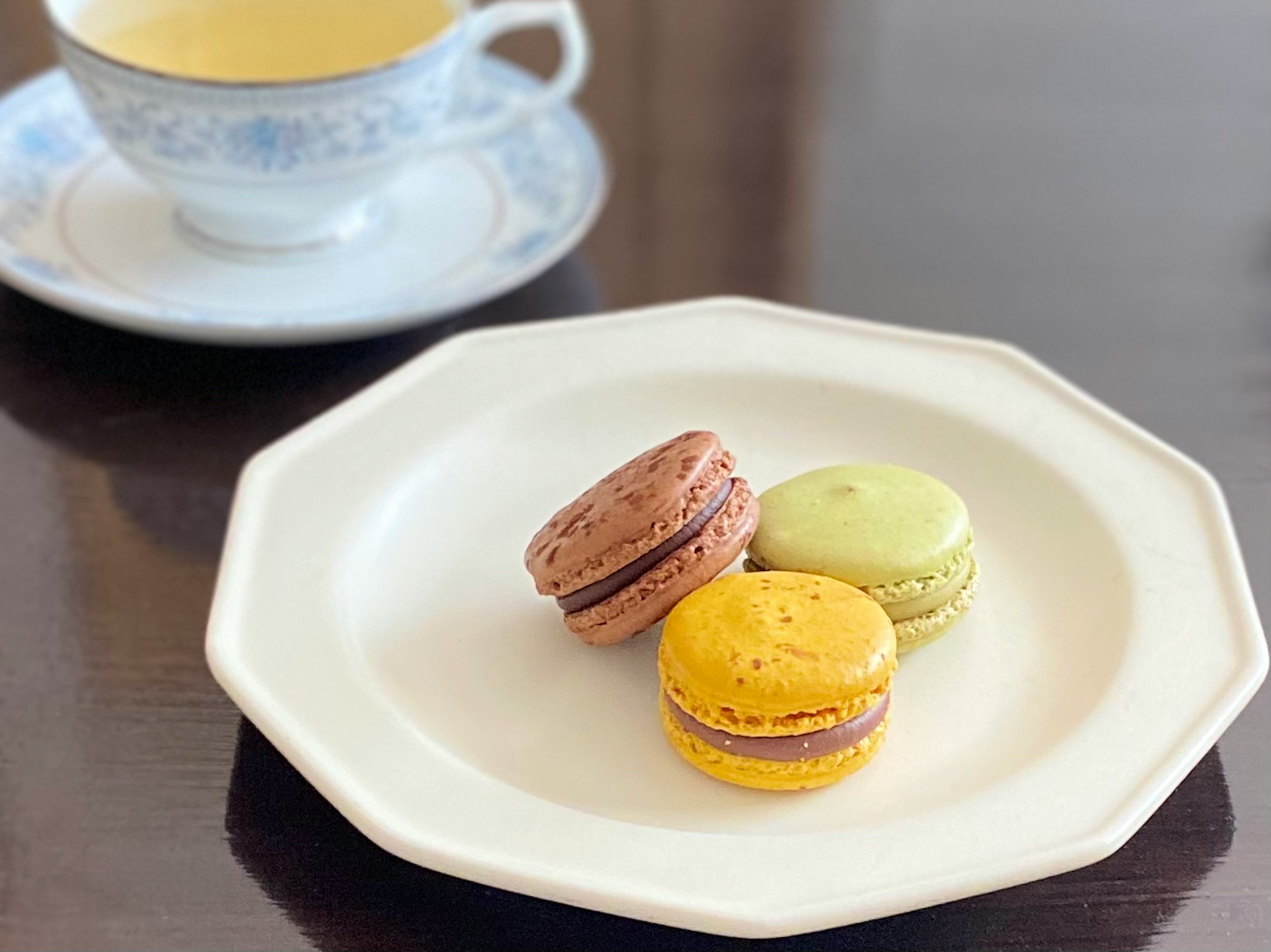 Three colorful macarons on a white plate with a teacup in the background