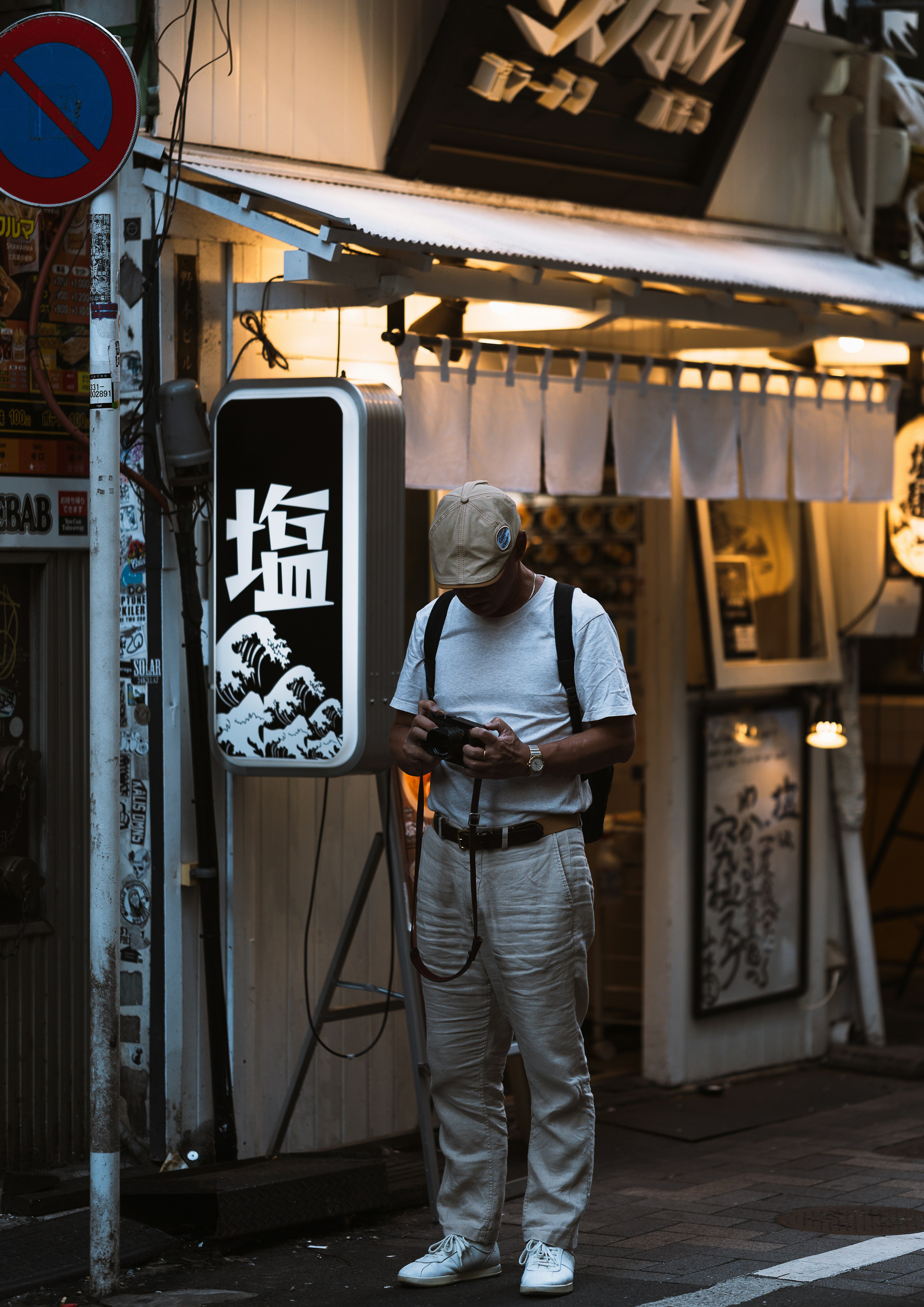 Un uomo che guarda il suo smartphone in un angolo di strada poco illuminato indossando una maglietta bianca e pantaloni vicino a un'insegna di ristorante giapponese