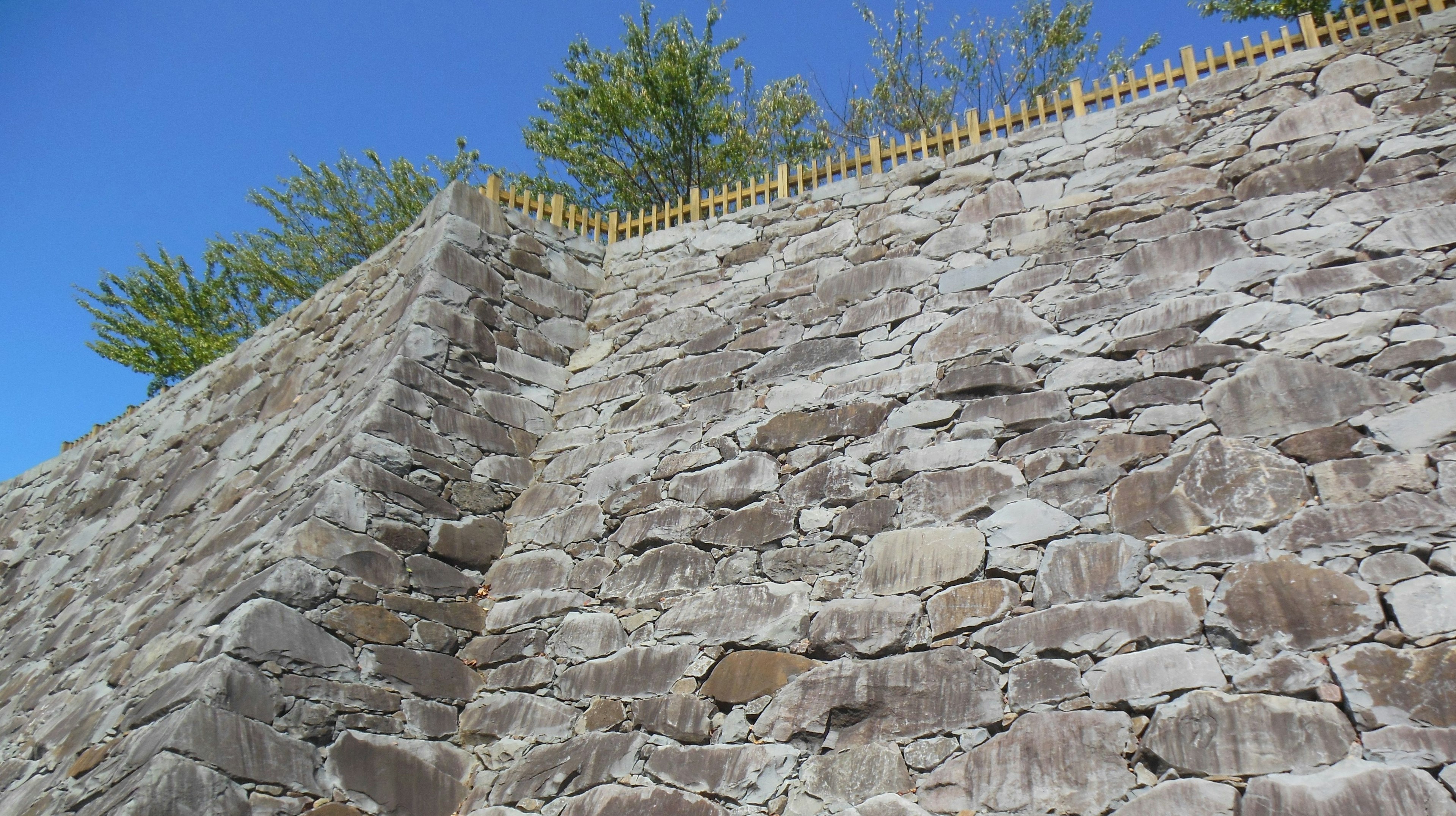 Muro de piedra con una valla dorada en la parte superior bajo un cielo azul y árboles verdes