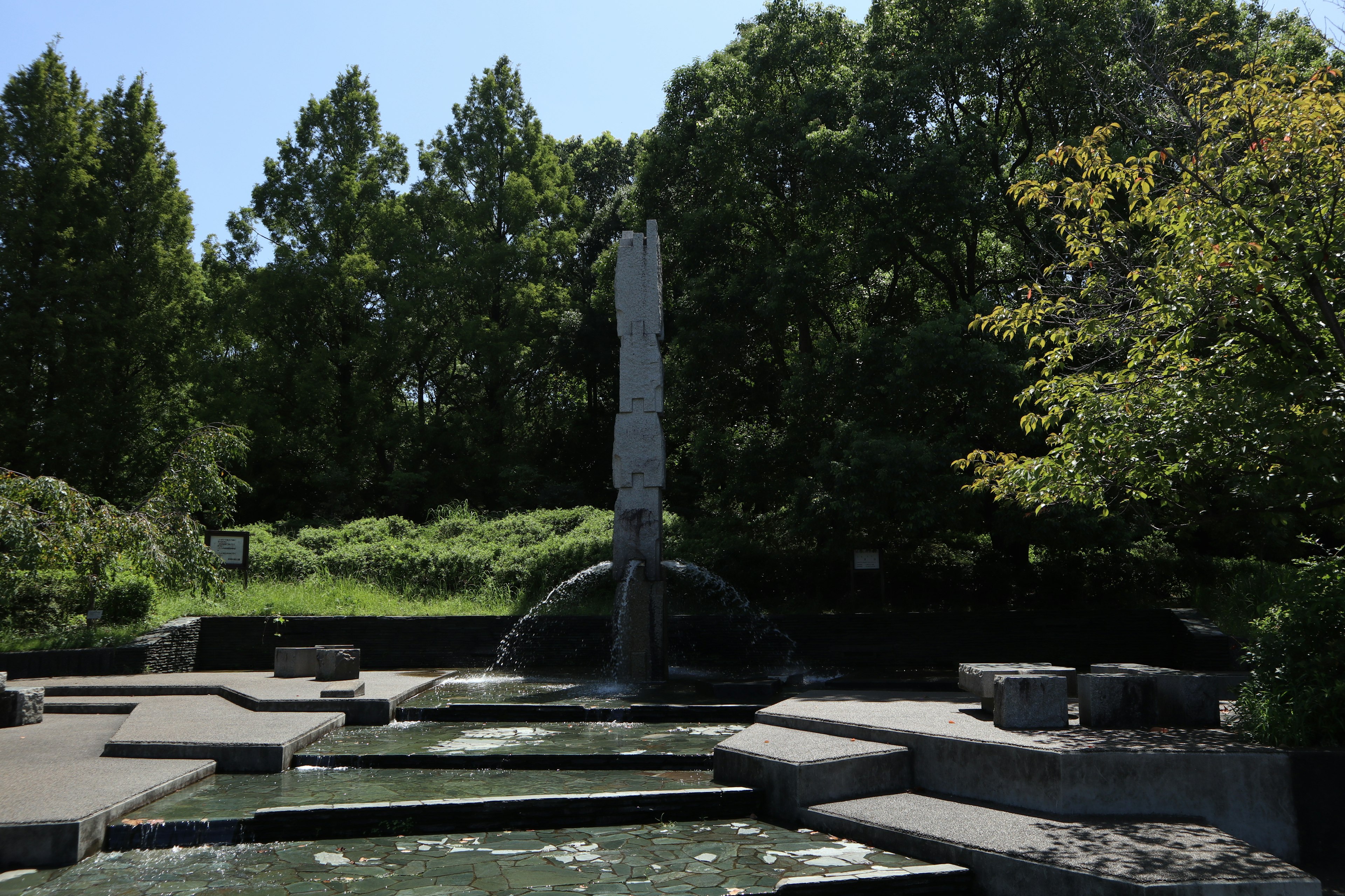 A tranquil park scene featuring a water fountain and a stone sculpture surrounded by greenery