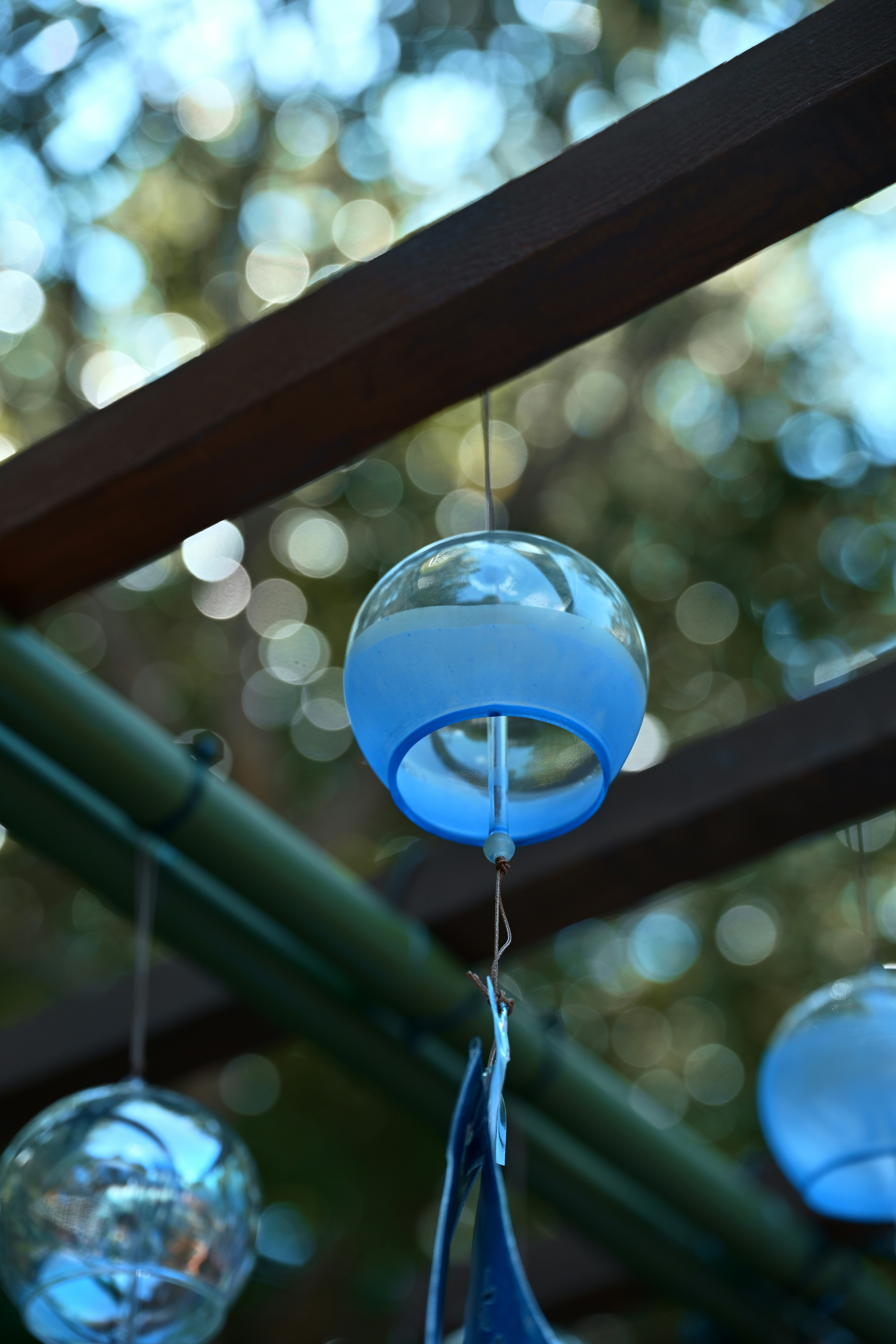 A blue glass wind chime hanging from a wooden beam with blurred green foliage in the background