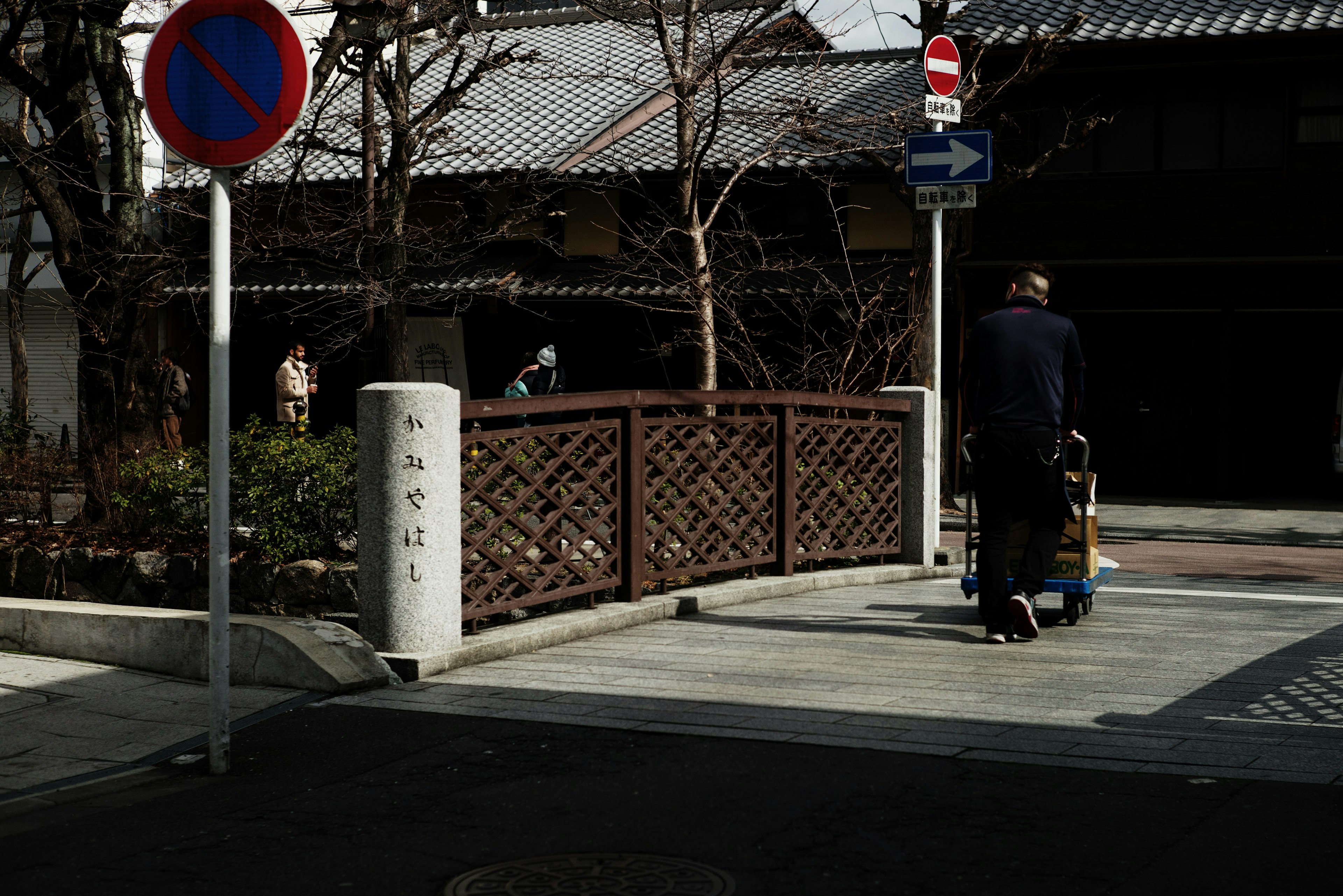 A person pushing a cart crosses the street with an old building and wooden fence in view