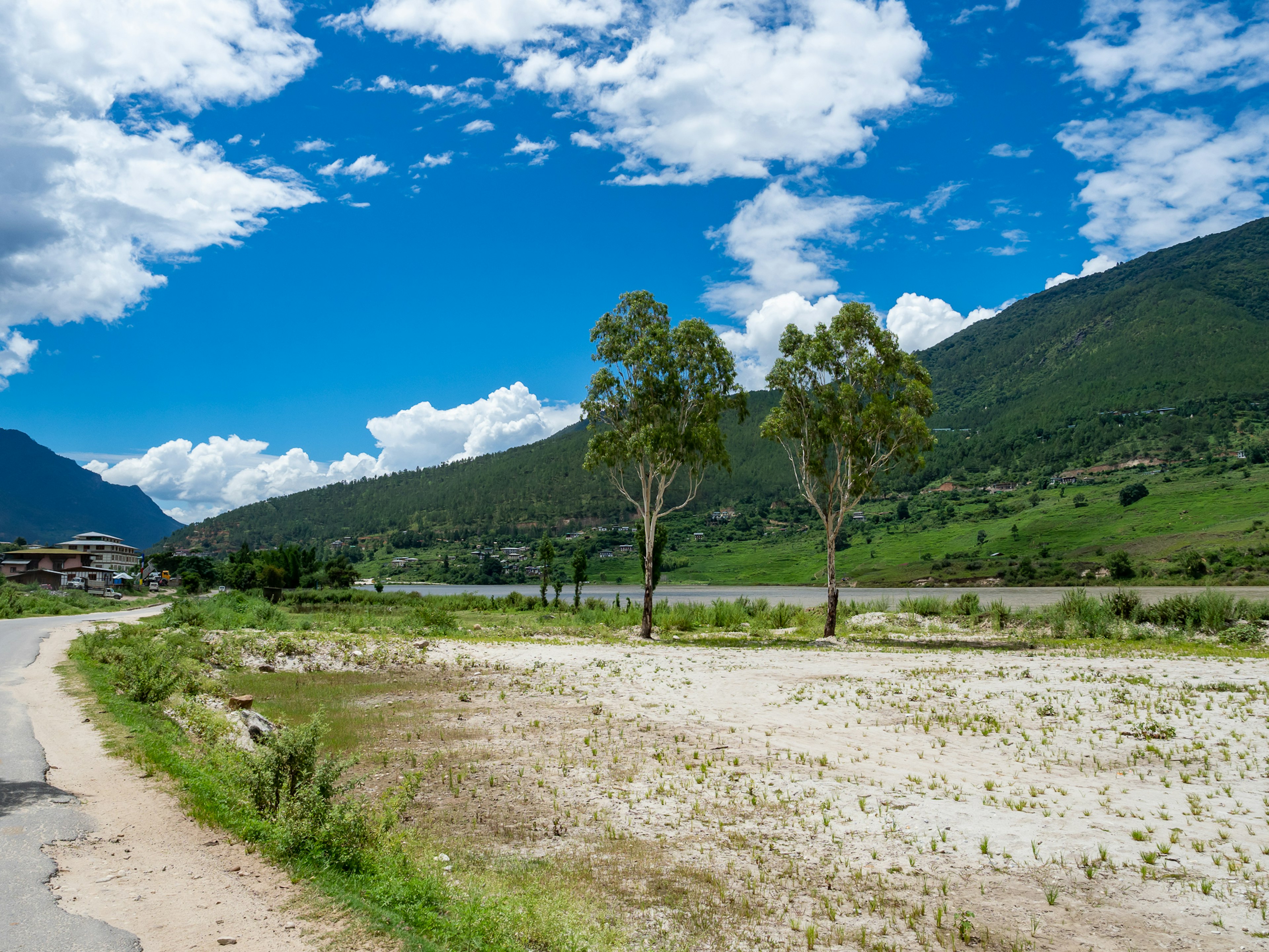 青い空と白い雲が広がる風景 緑の山々と川が見える 砂利の道と木々が点在する