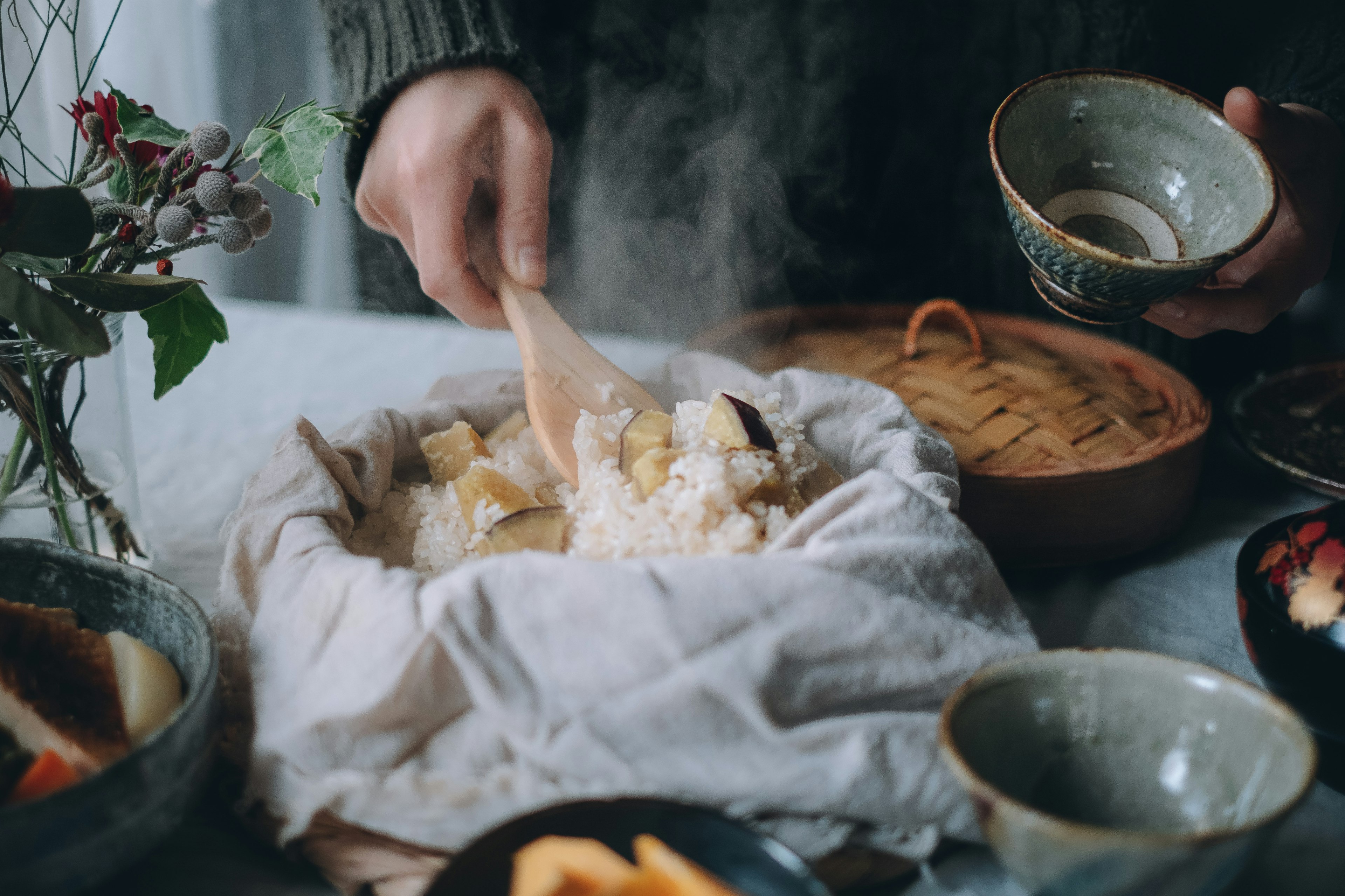 Une main remuant du riz d'un cuiseur à vapeur avec une cuillère en bois entourée de bols et de nourriture