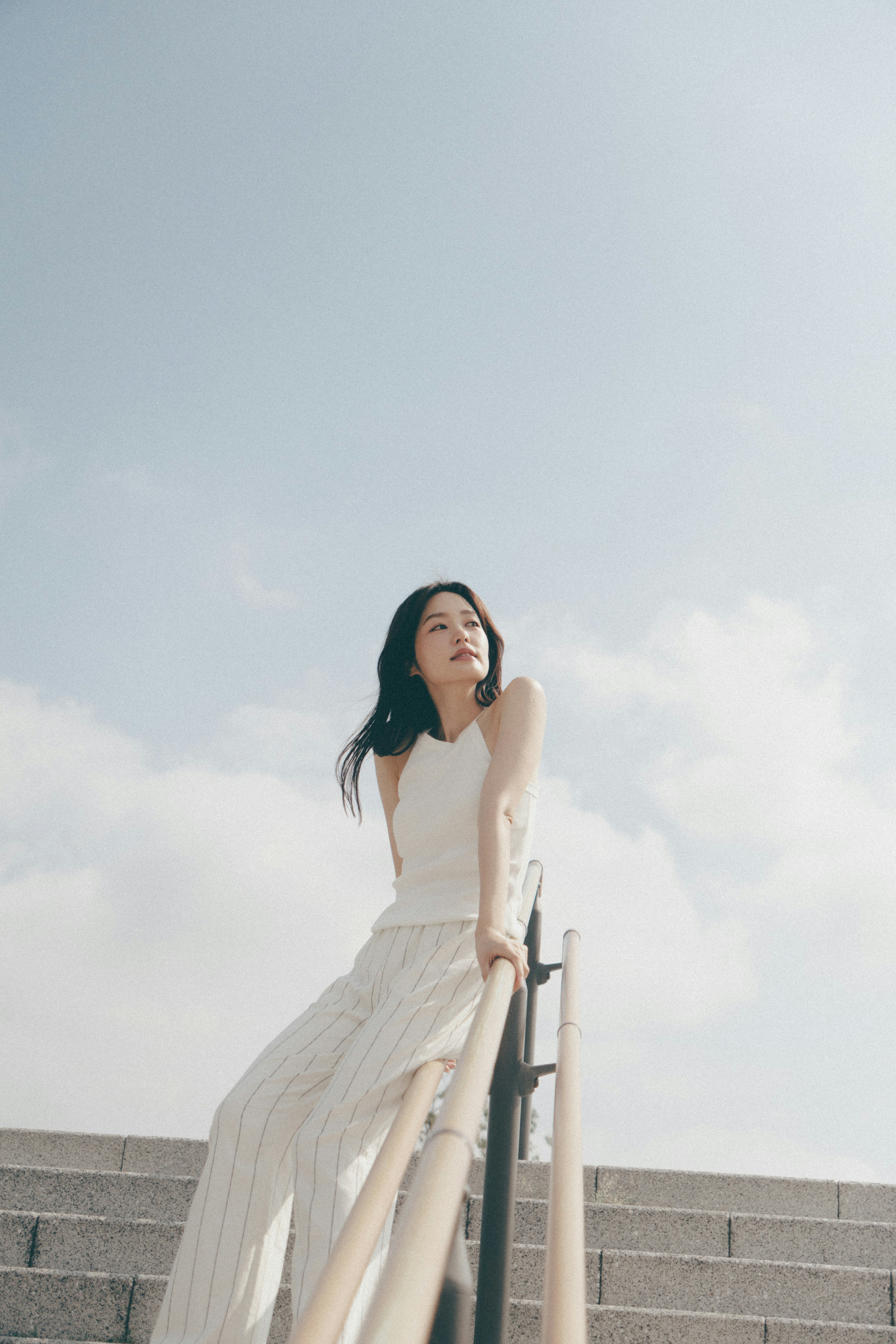 Porträt einer Frau, die auf Treppen unter einem blauen Himmel sitzt, in einem weißen Outfit mit langen Haaren, die im Wind wehen