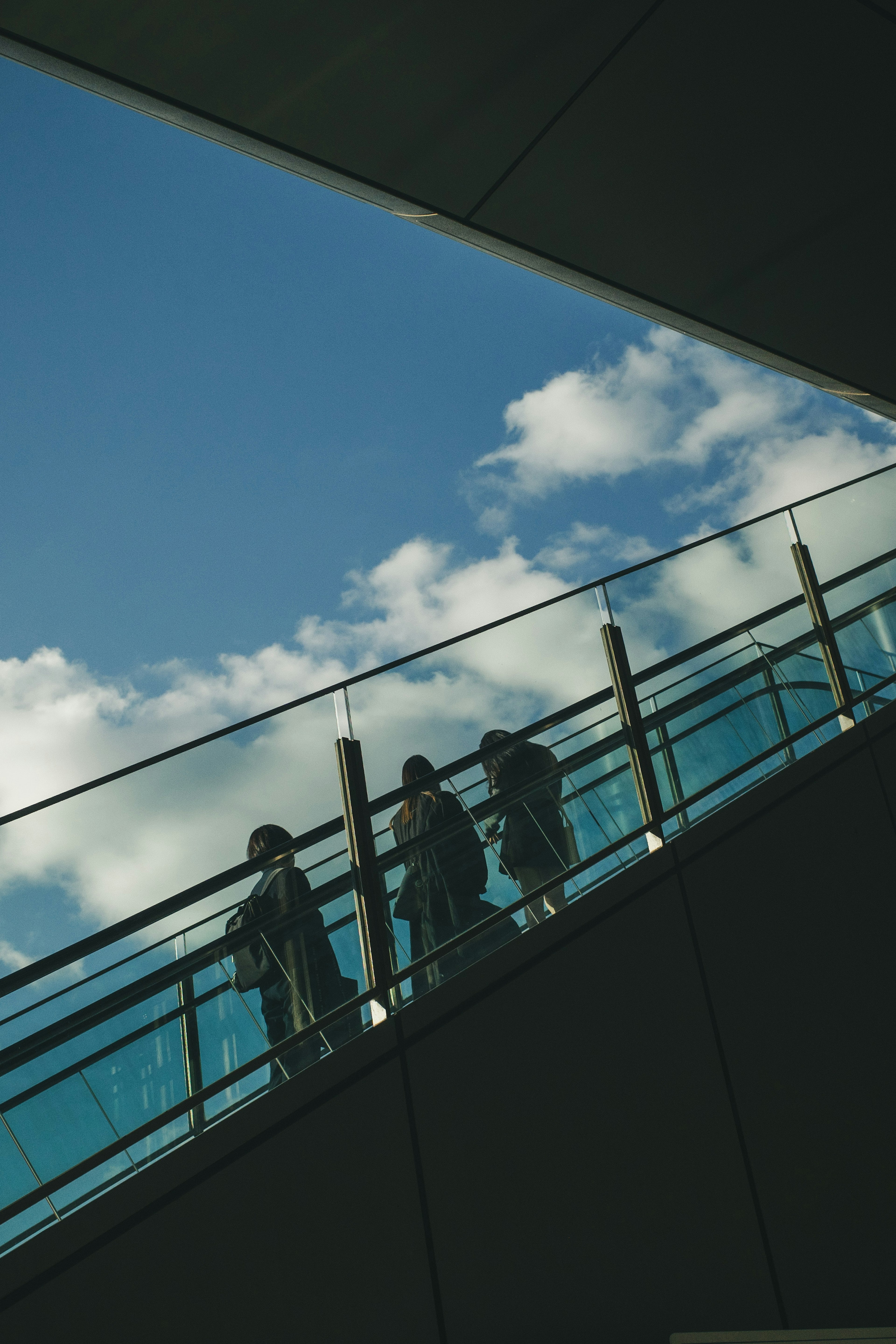 Silhouettes de personnes marchant le long d'une rampe en verre sous un ciel bleu et des nuages