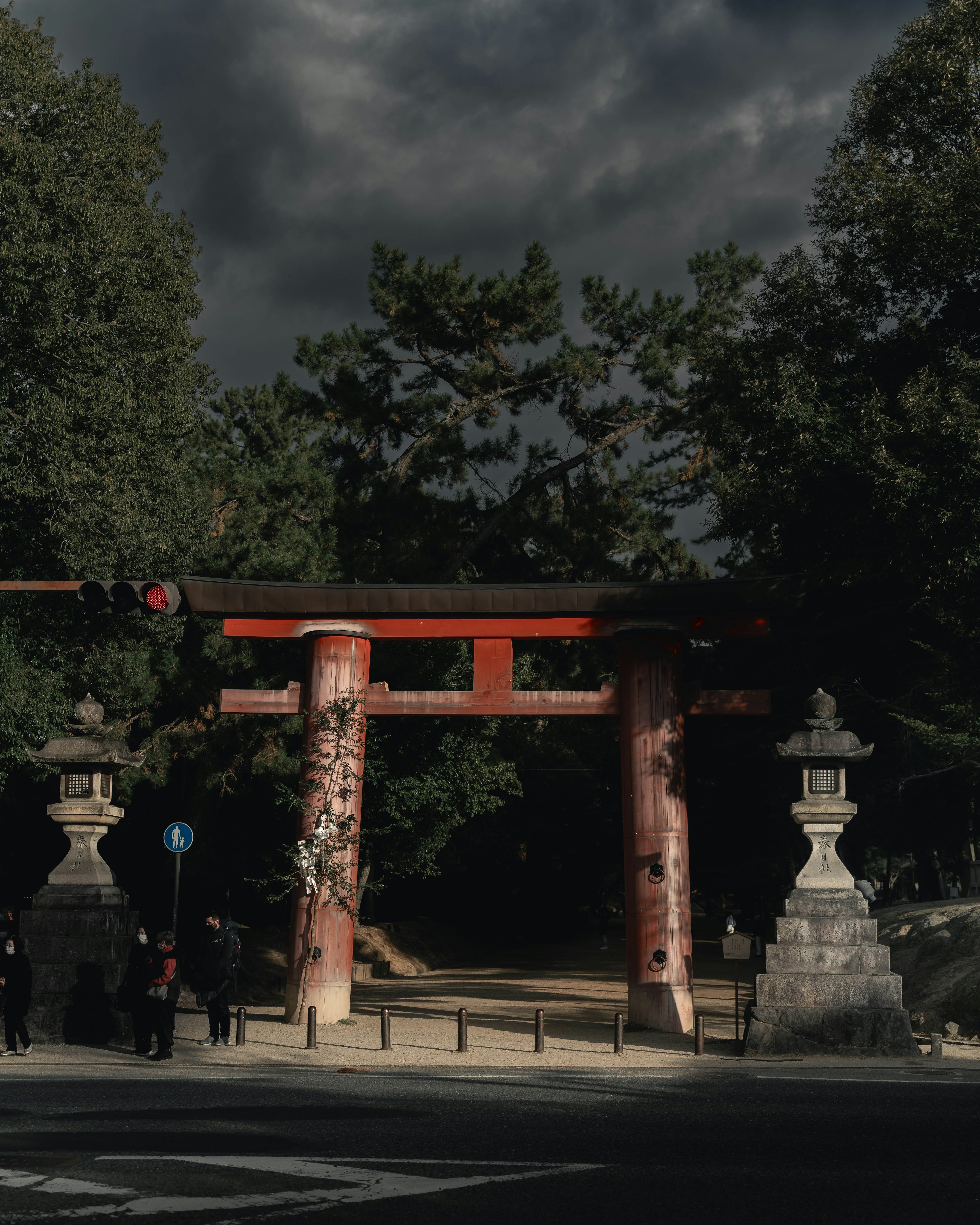 Entrance of a shrine featuring a red torii gate and stone lanterns dark clouds and trees in the background