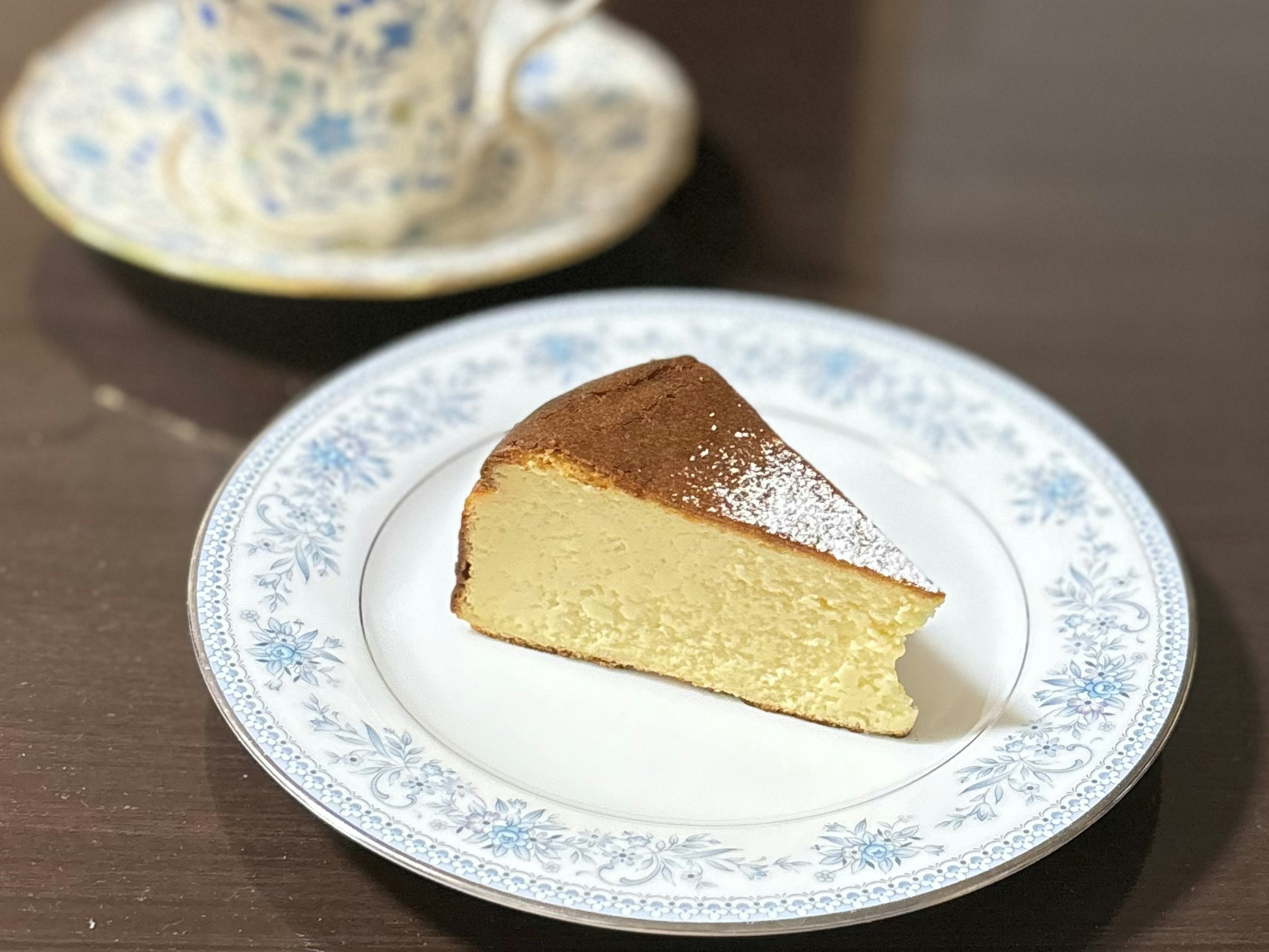 Ein Stück Biskuitkuchen auf einem dekorativen Blumenteller mit einer Tasse im Hintergrund