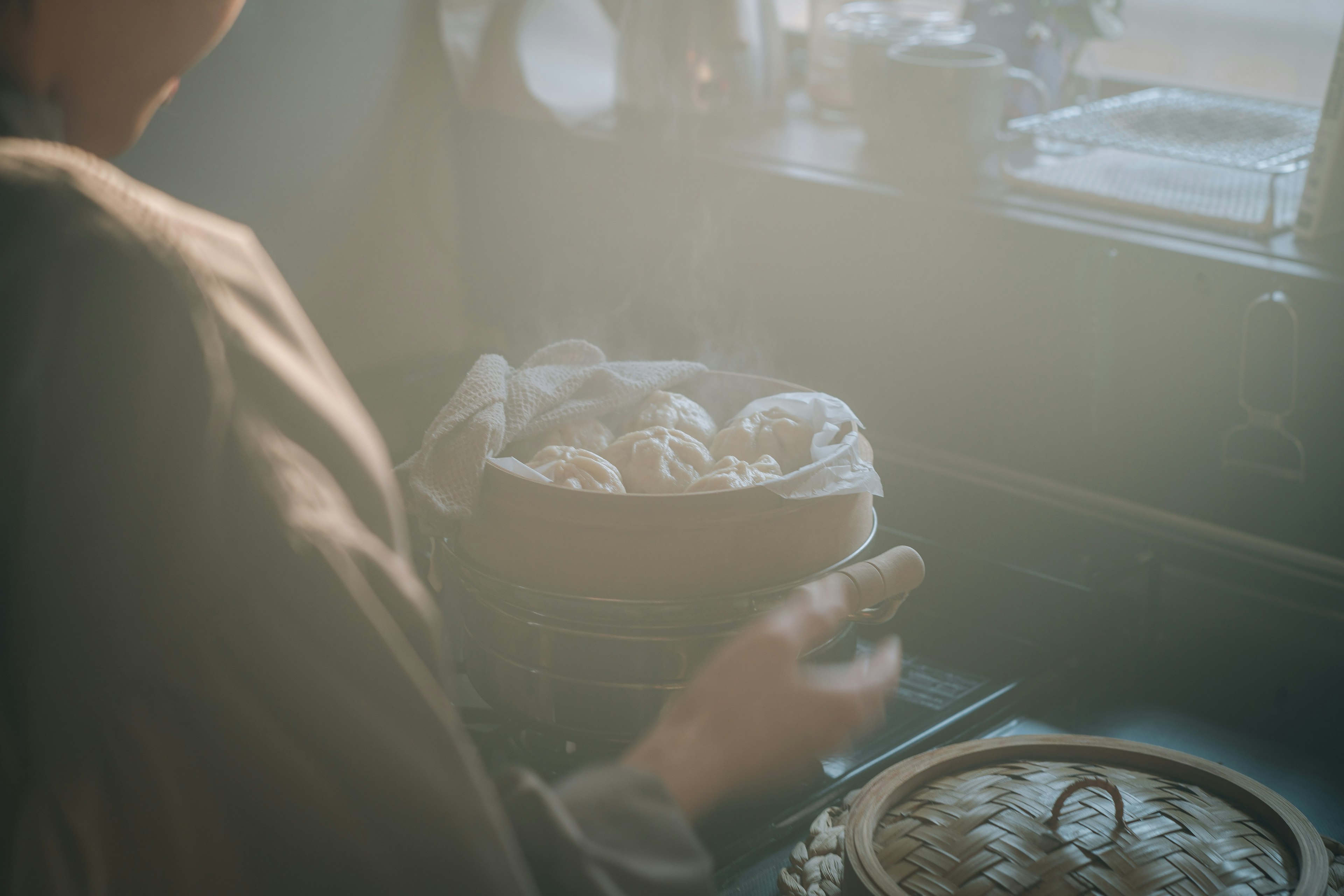 温かい蒸し料理を準備している人物の後ろ姿、窓からの柔らかい光が差し込むキッチン