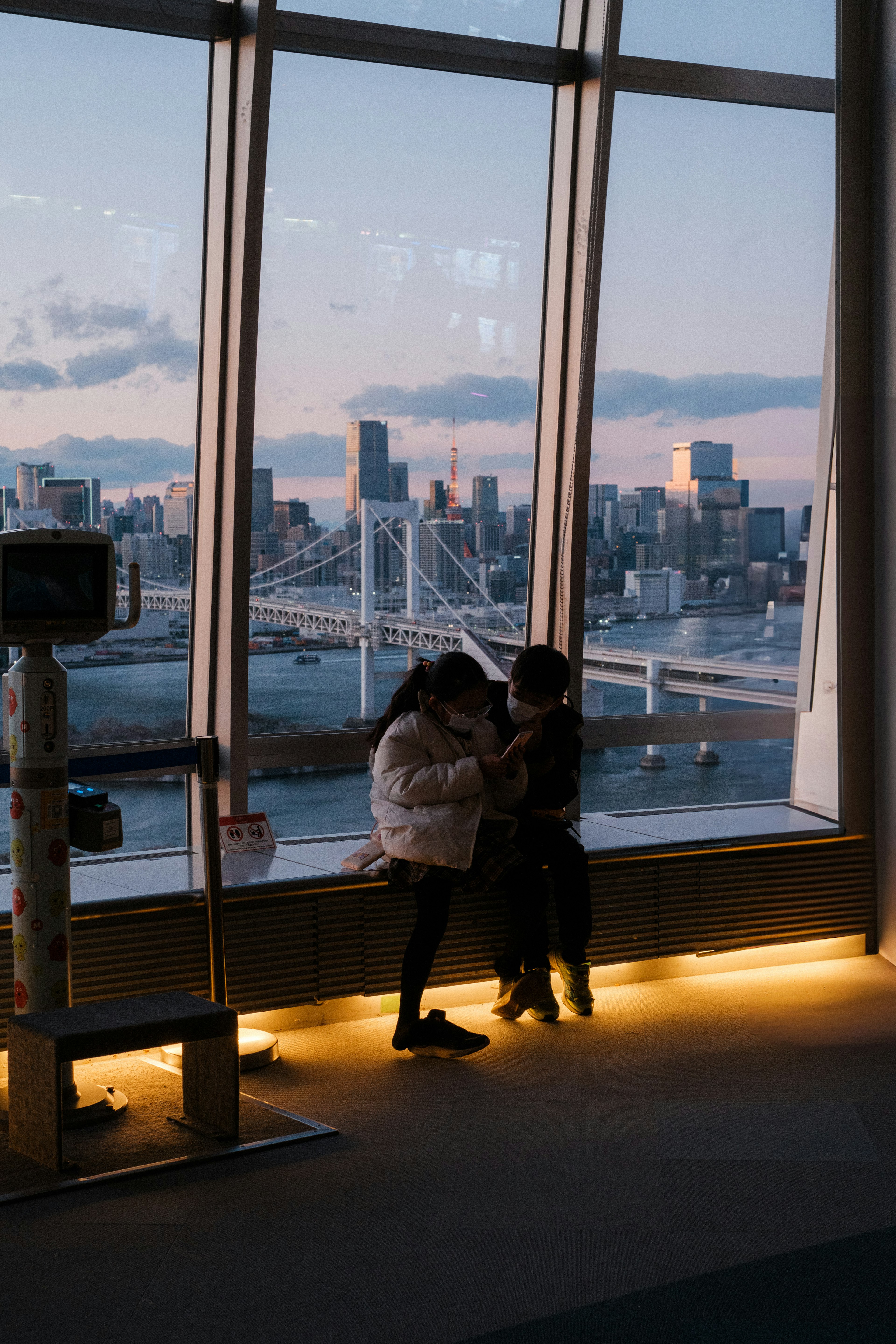 Dos mujeres compartiendo un momento junto a una gran ventana con vista a la bahía de Tokio al atardecer