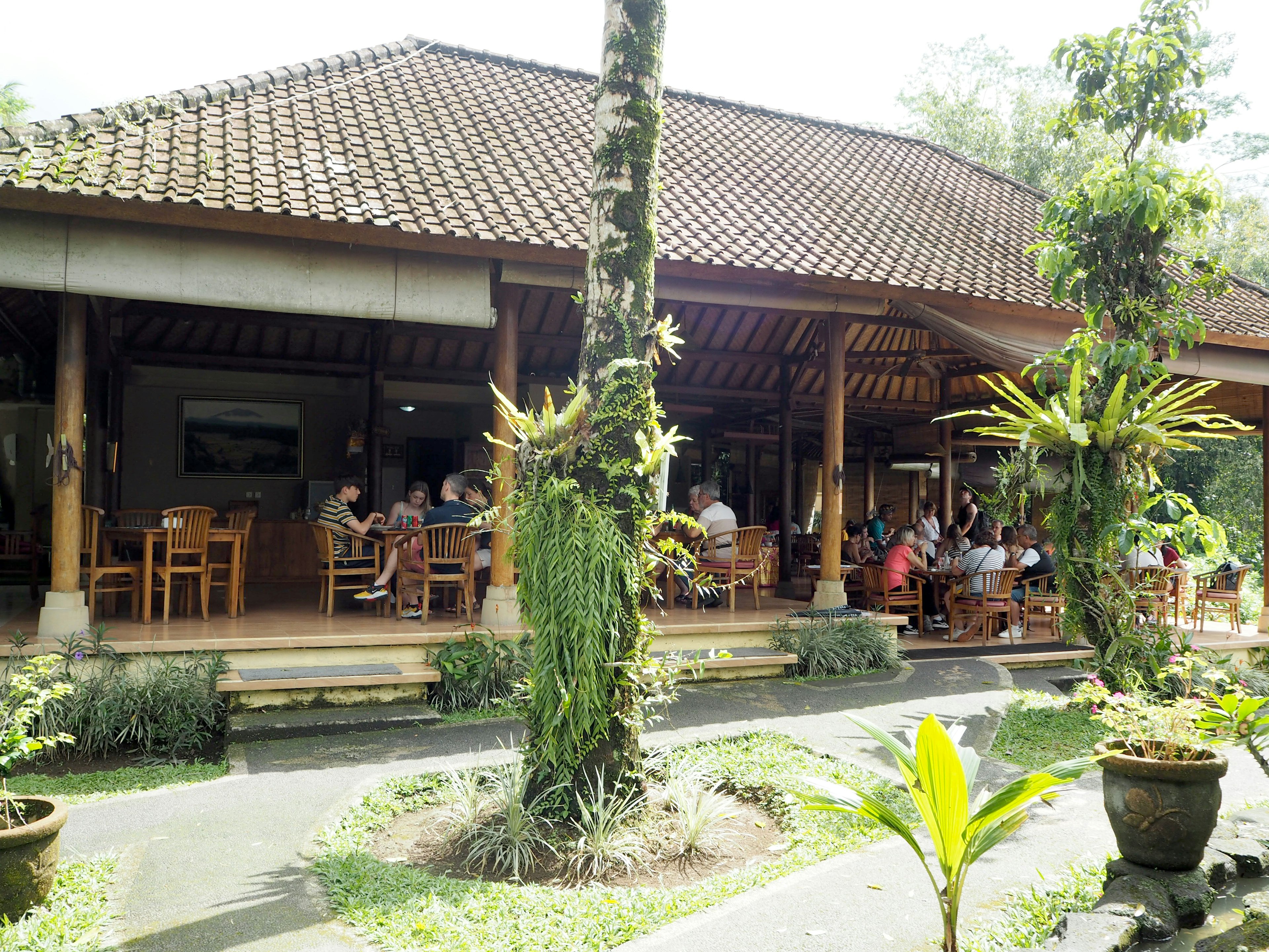 Außenansicht eines Restaurants in Bali mit üppigem Grün und Holztischen und -stühlen