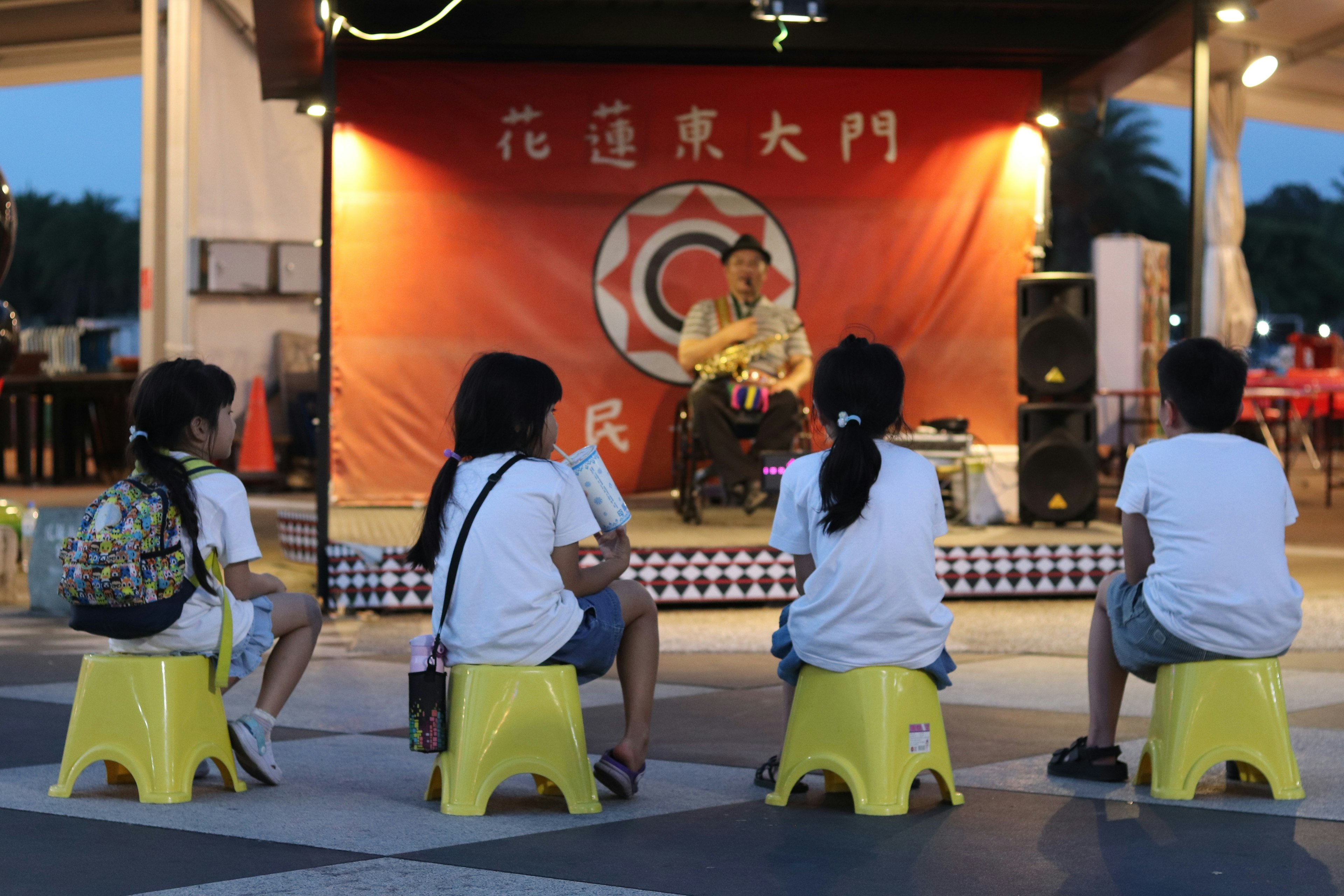 子供たちが黄色い椅子に座ってステージを見ている夜の風景