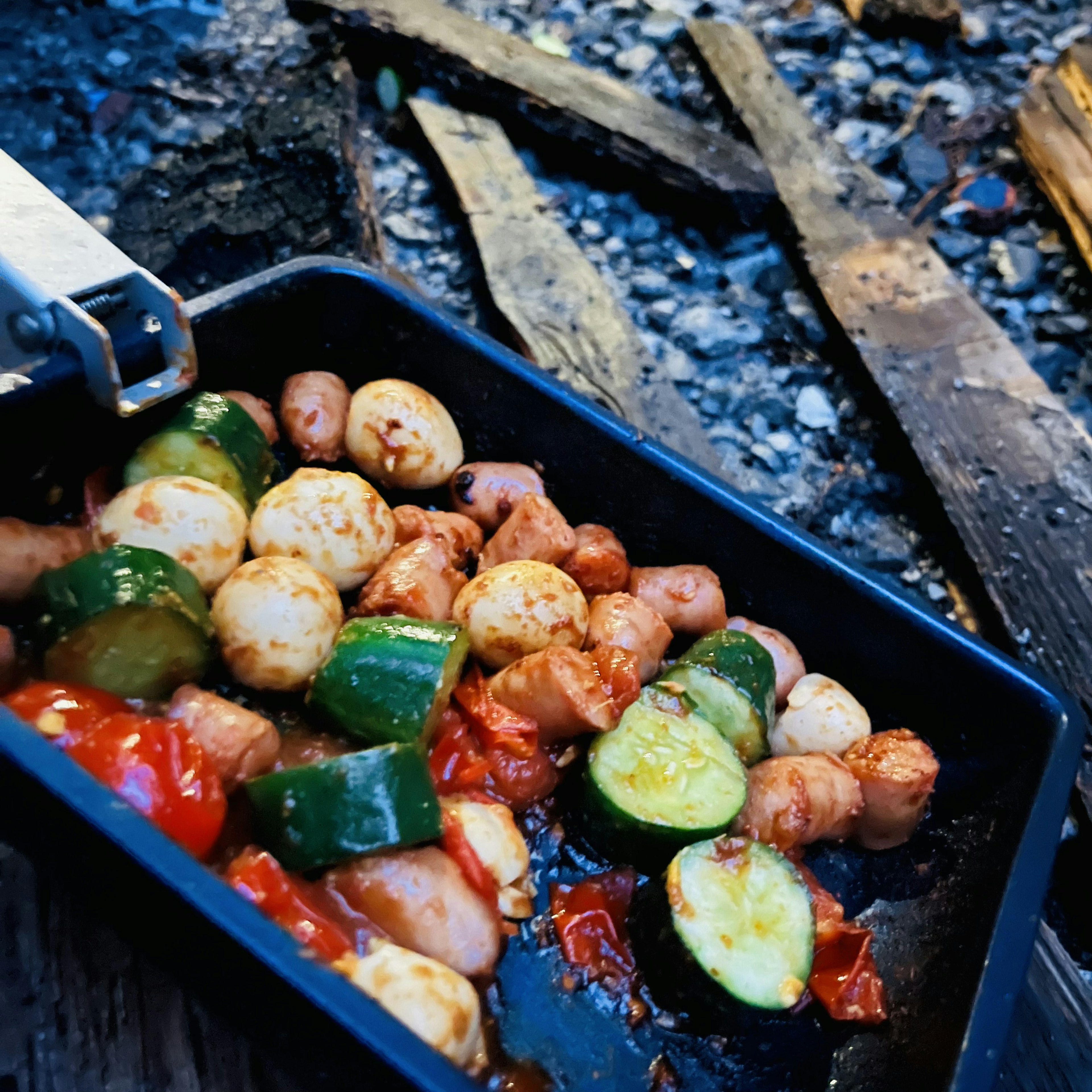 Une poêle de légumes et saucisses cuits près d'un feu de camp