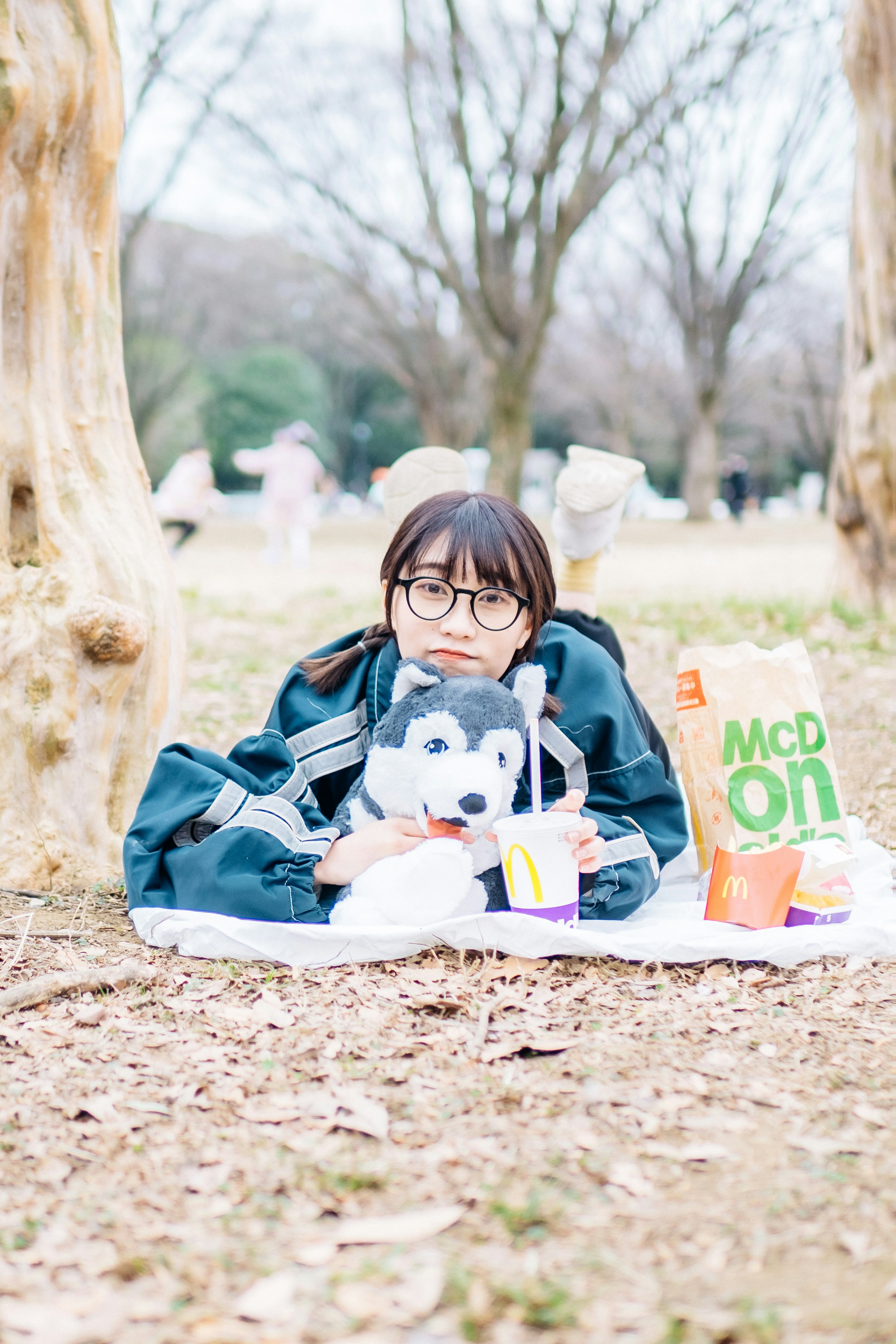 A girl sitting on a blanket in a park with a stuffed dog beside her food and drink items