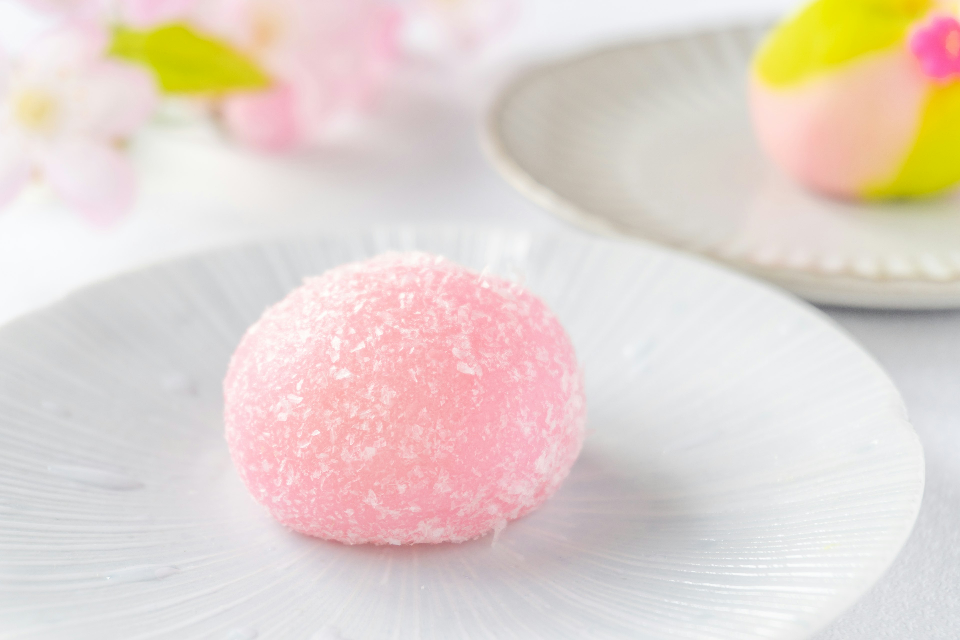 A soft pink Japanese sweet placed on a white plate with cherry blossom petals in the background