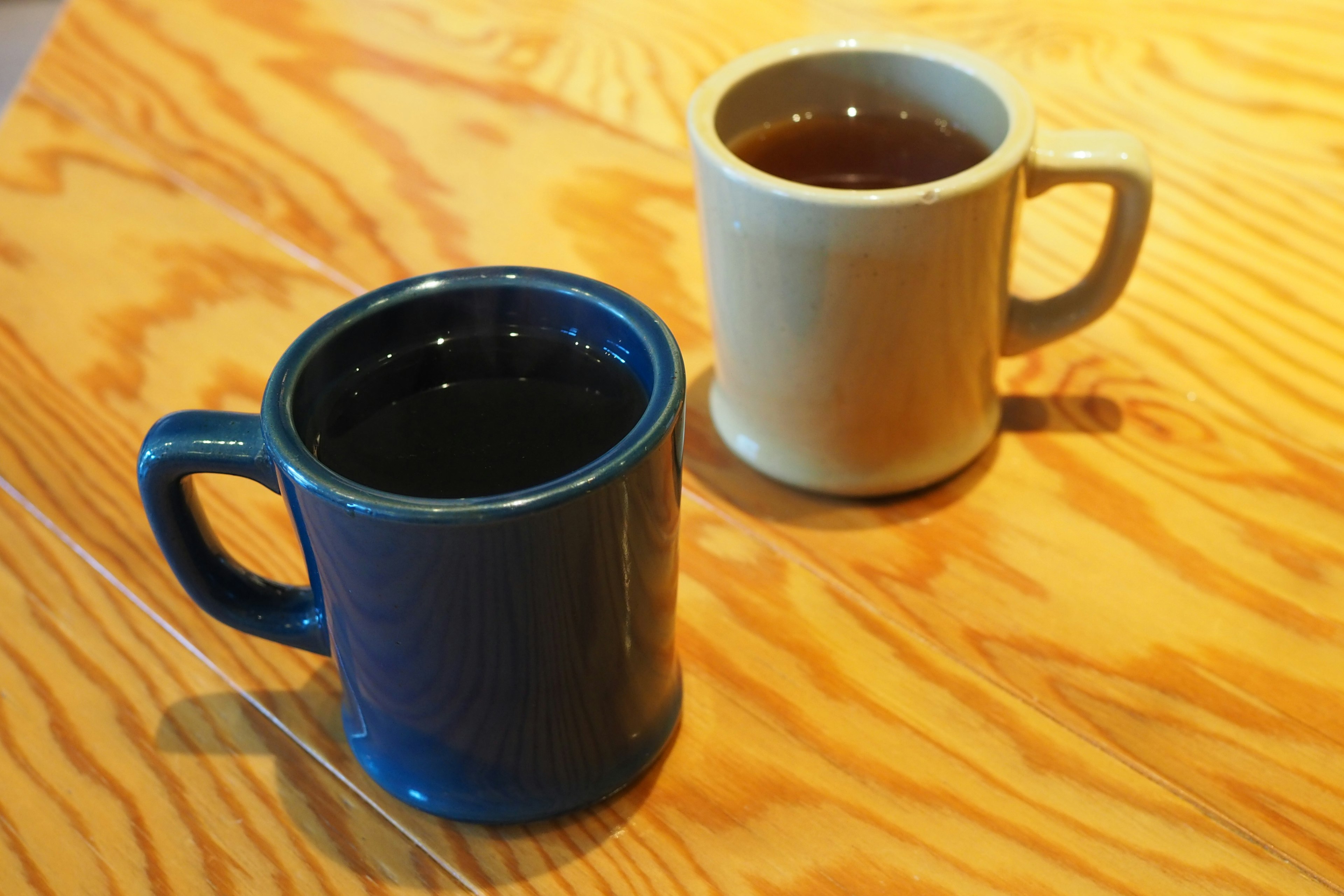 A blue mug and a cream-colored mug filled with tea placed on a wooden table