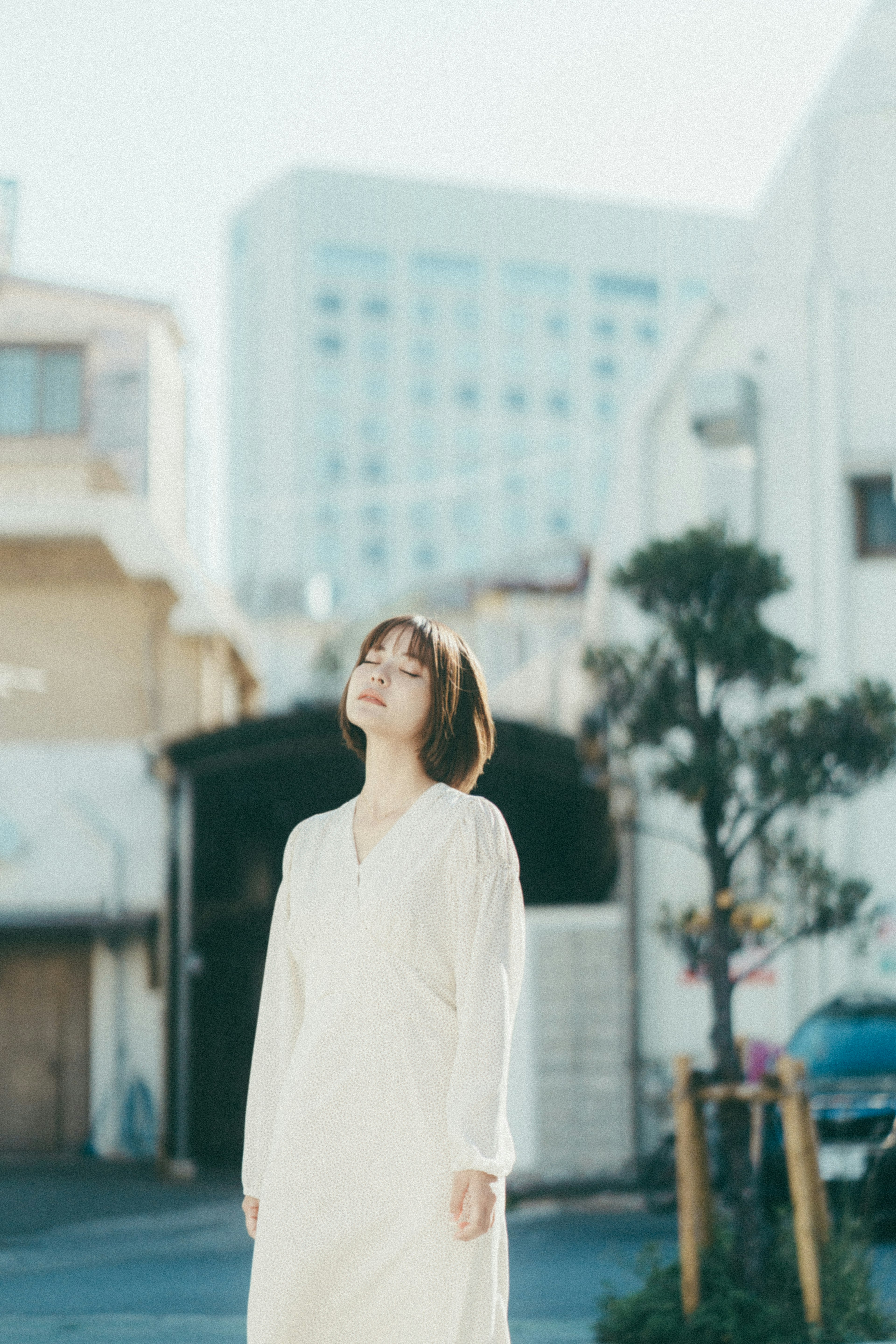 하얀 드레스를 입은 여성이 밝은 햇빛 속에 서 있고 배경에는 도시 건물이 있다