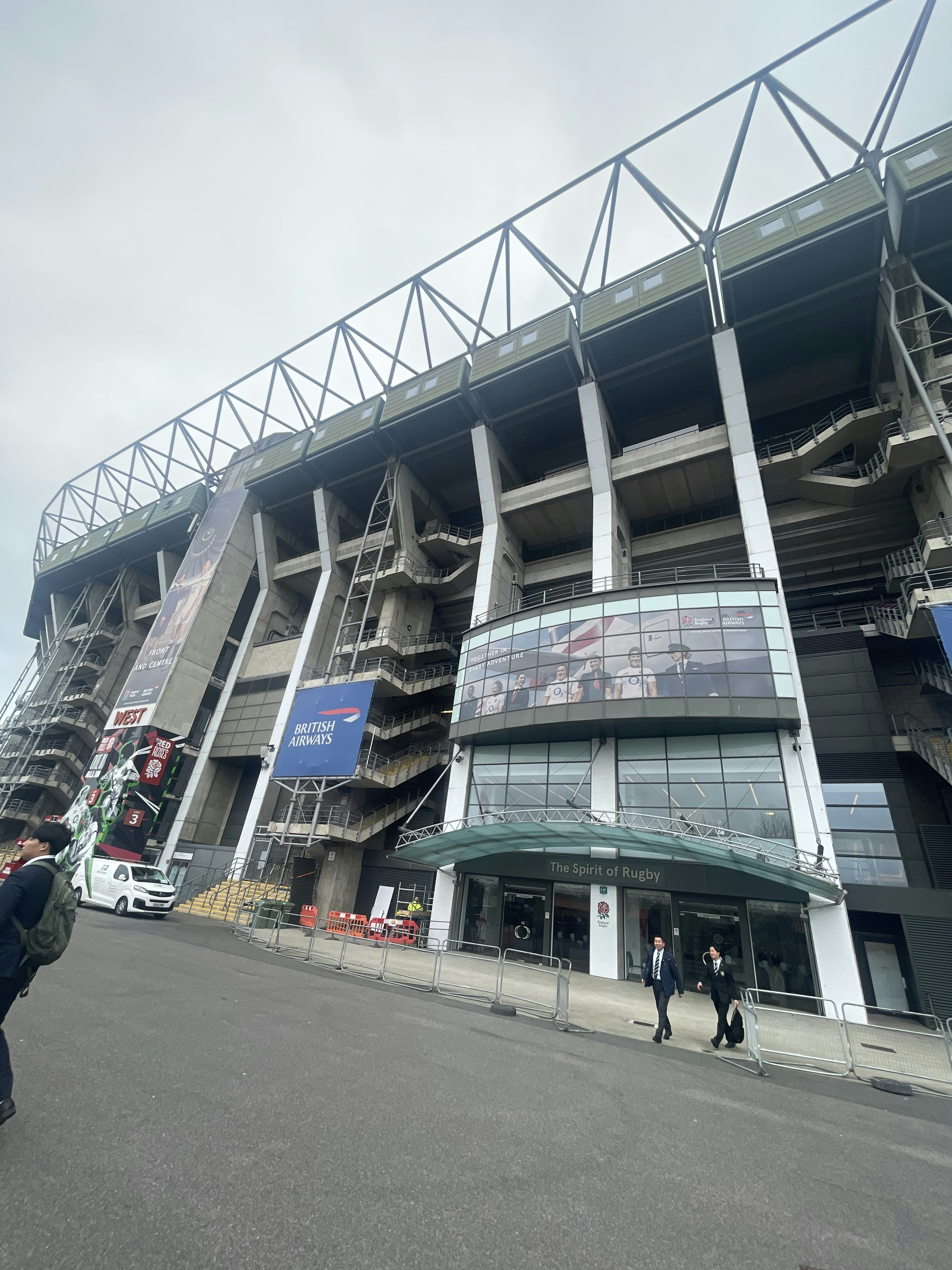 Gambar yang menunjukkan eksterior stadion desain arsitektur modern fasad kaca besar pintu masuk penonton terlihat