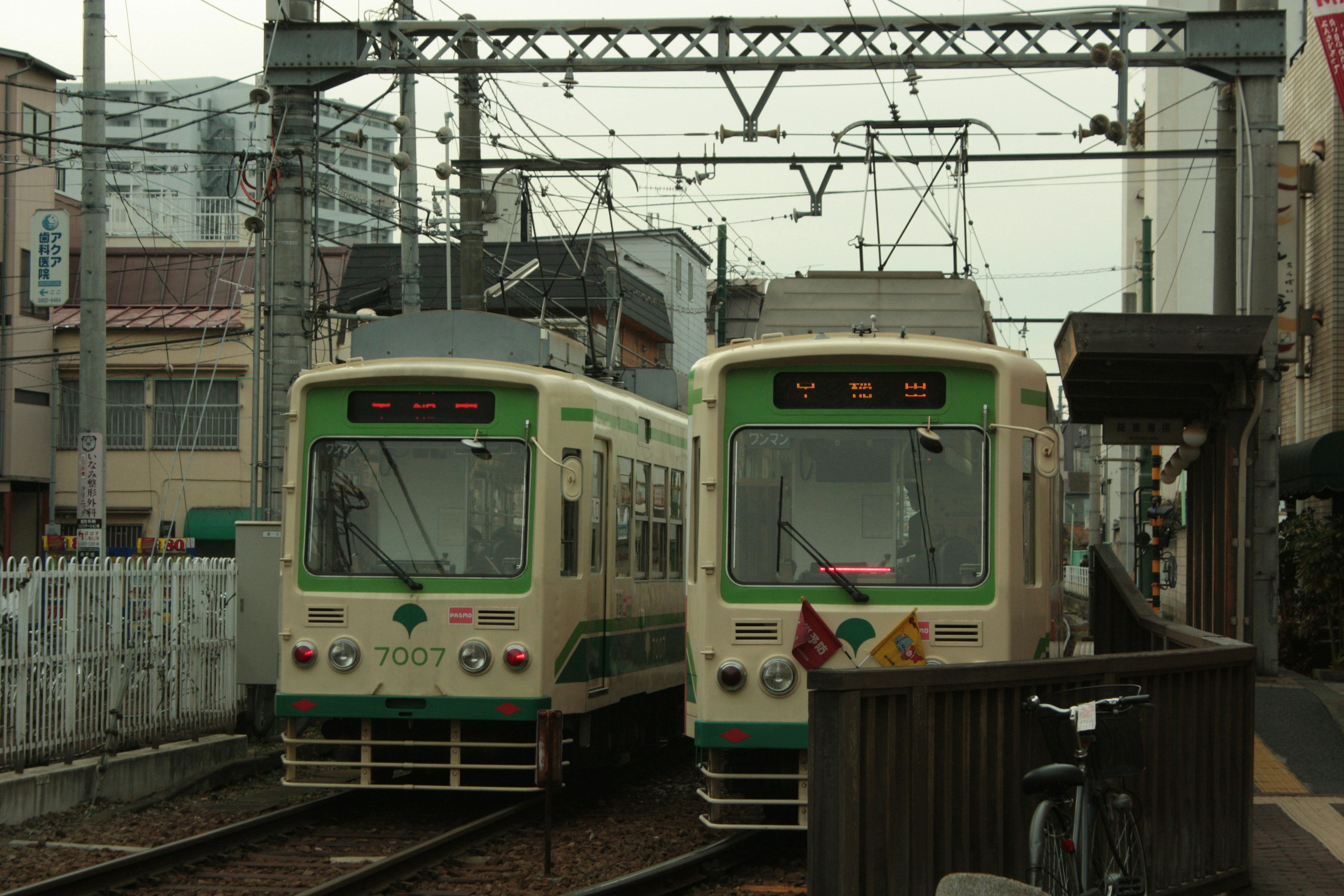 Dos tranvías están estacionados en una estación de tren con líneas aéreas y edificios urbanos de fondo