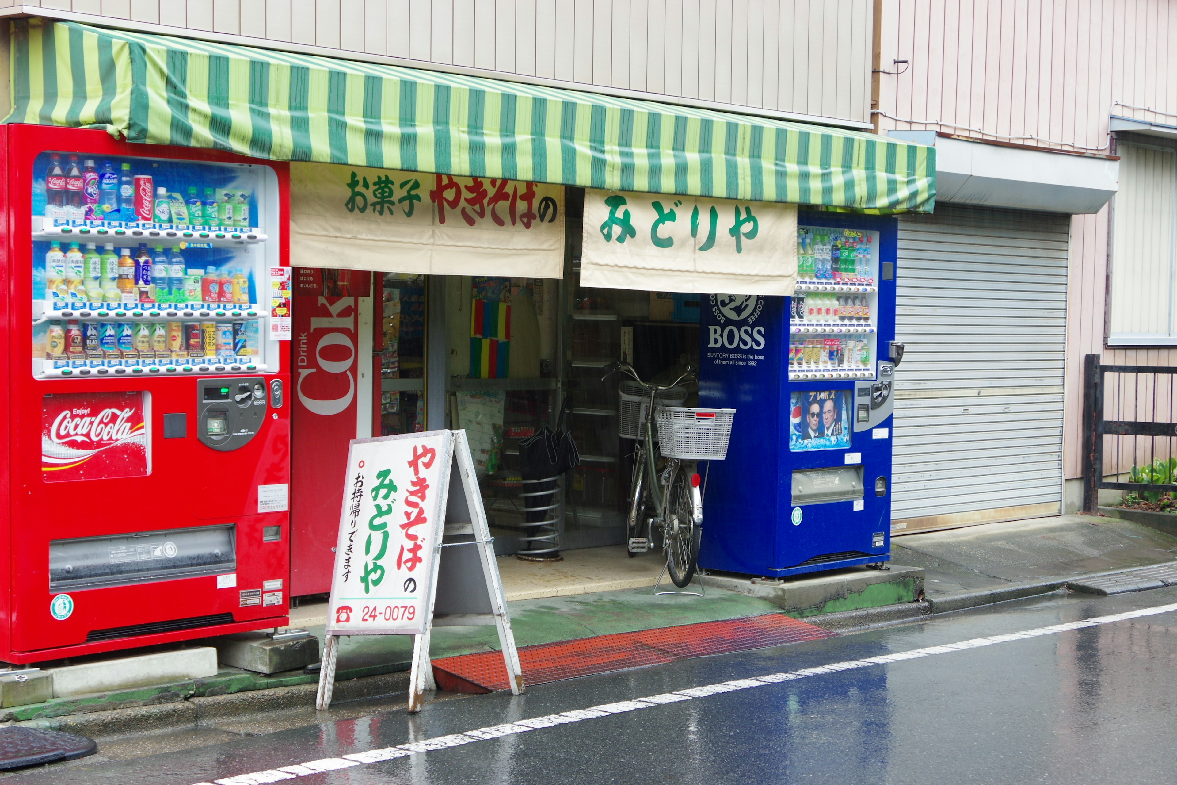 Extérieur d'un petit magasin avec des distributeurs automatiques devant Route mouillée et auvent rayé vert