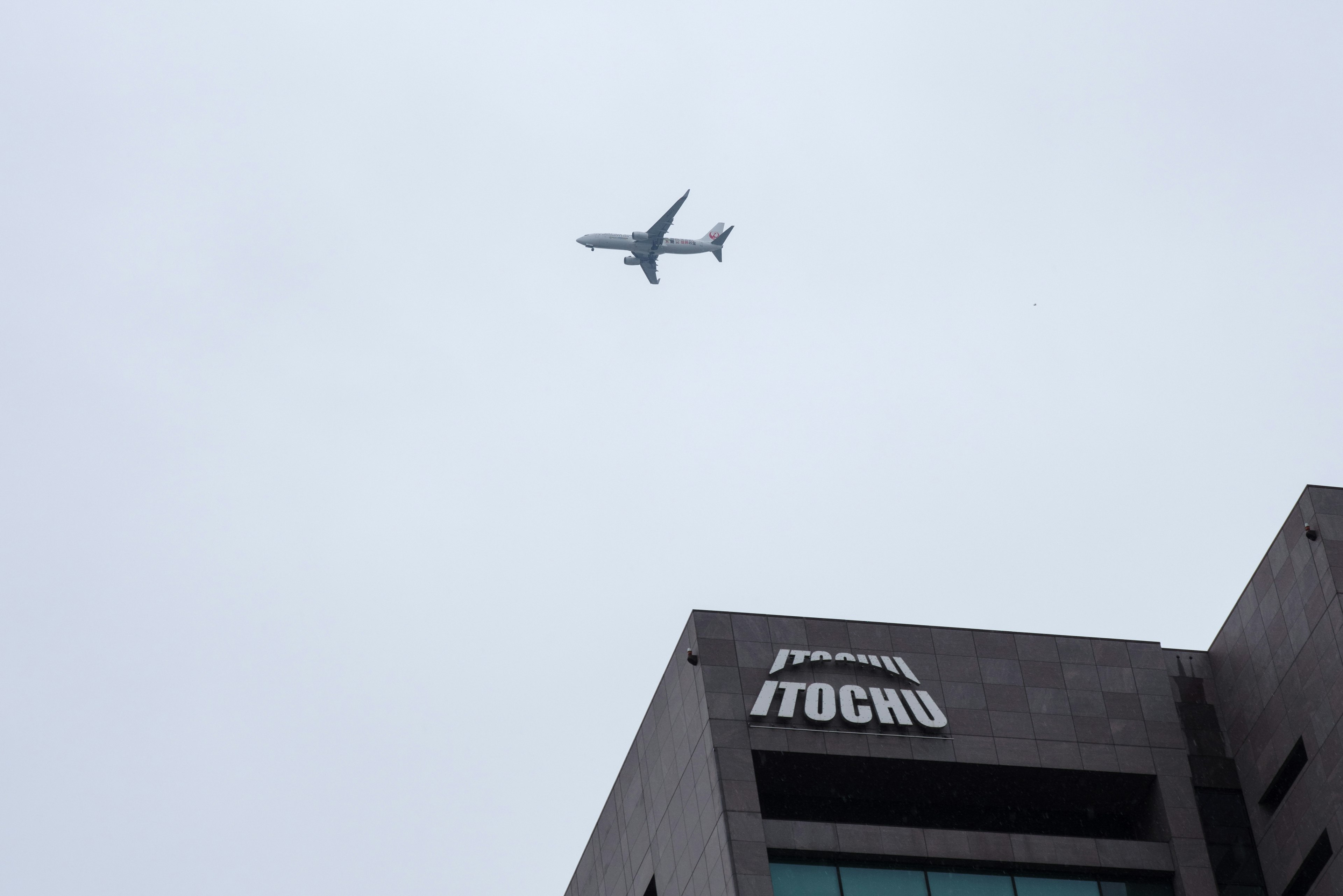 An airplane flying in a cloudy sky above a building with the ITOCHU sign