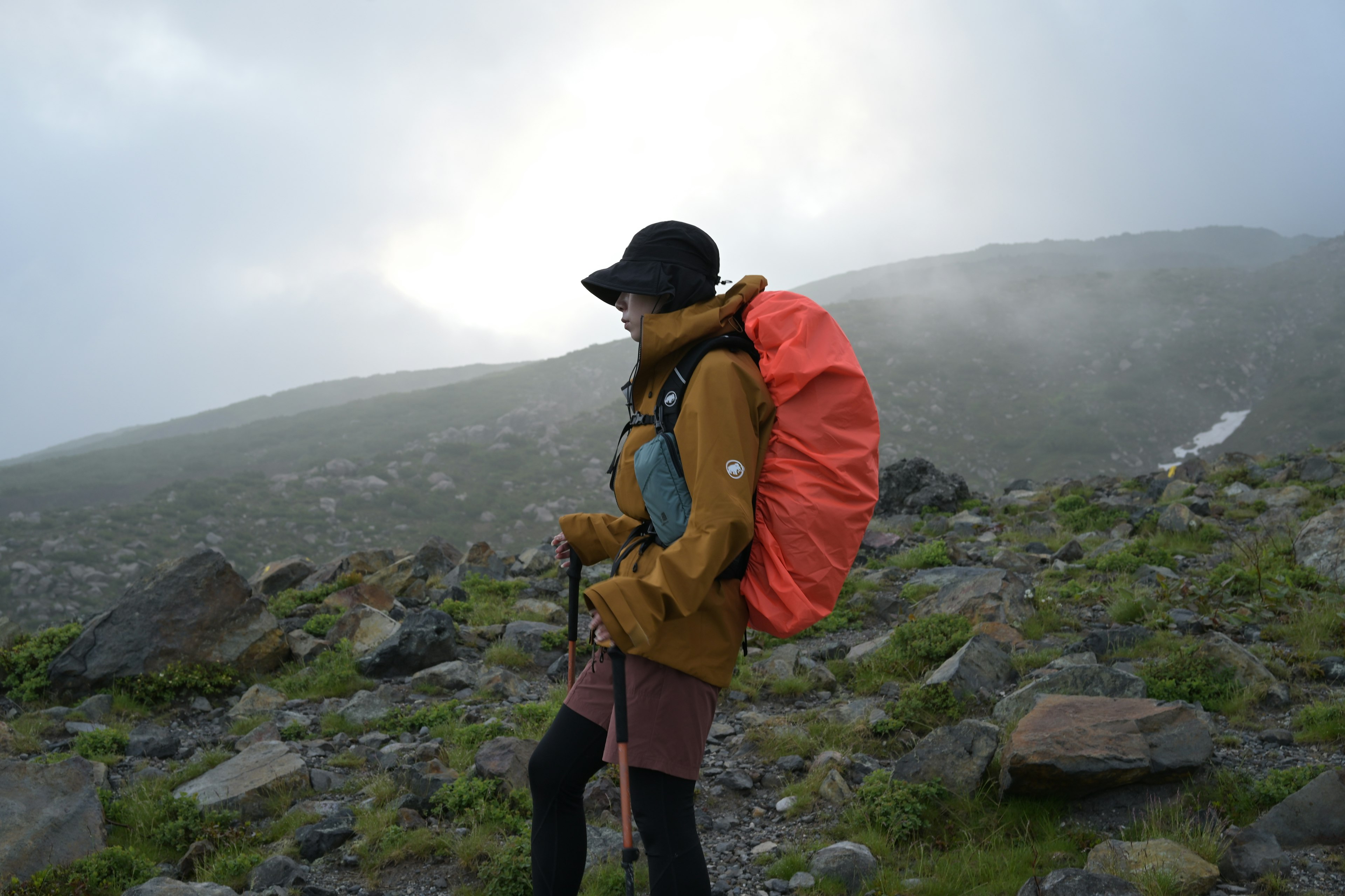 A woman hiking in fog wearing an orange backpack against a rocky landscape