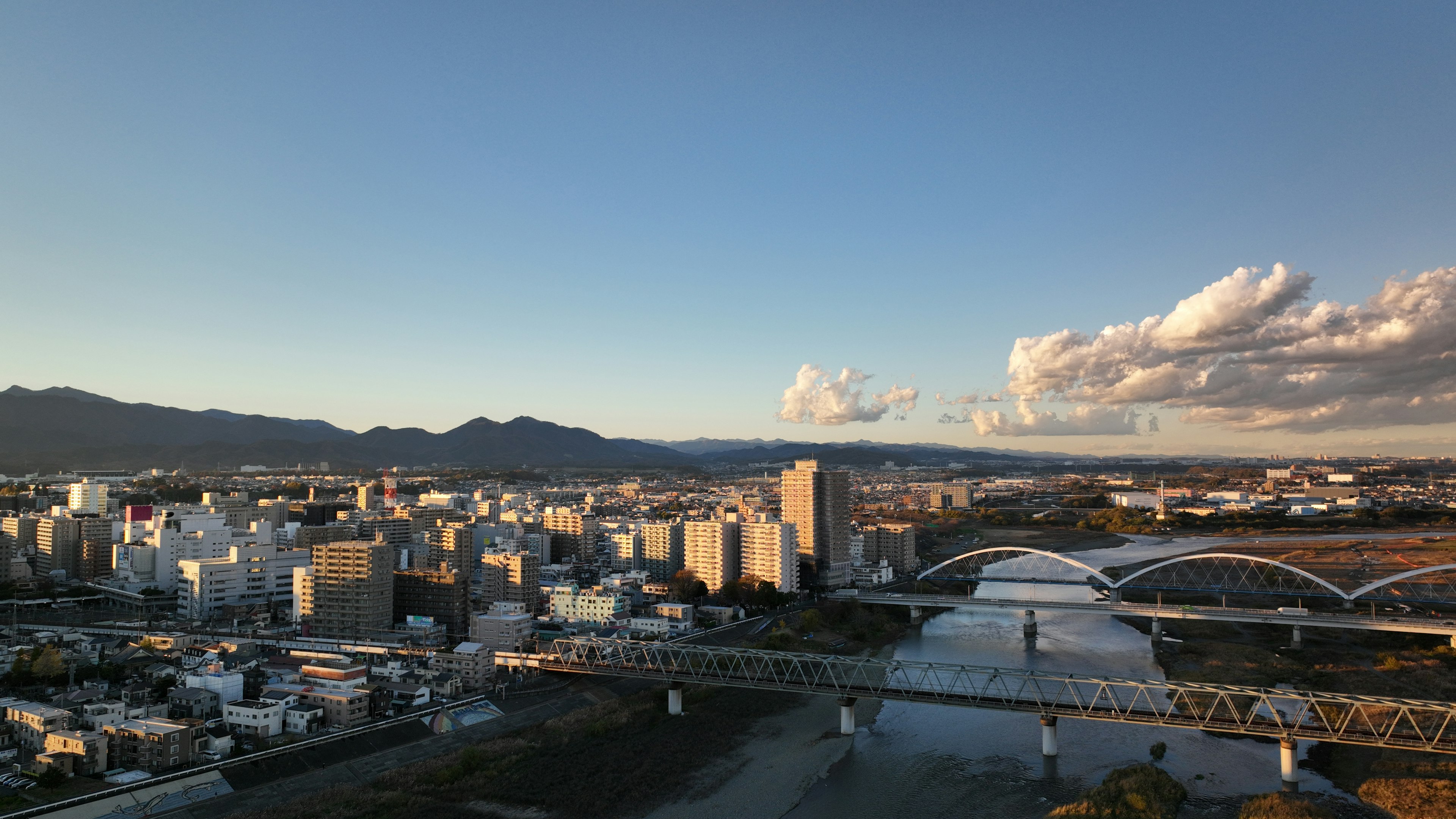 高層ビルと橋が並ぶ都市の風景で、川が流れ、青空に白い雲が浮かぶ