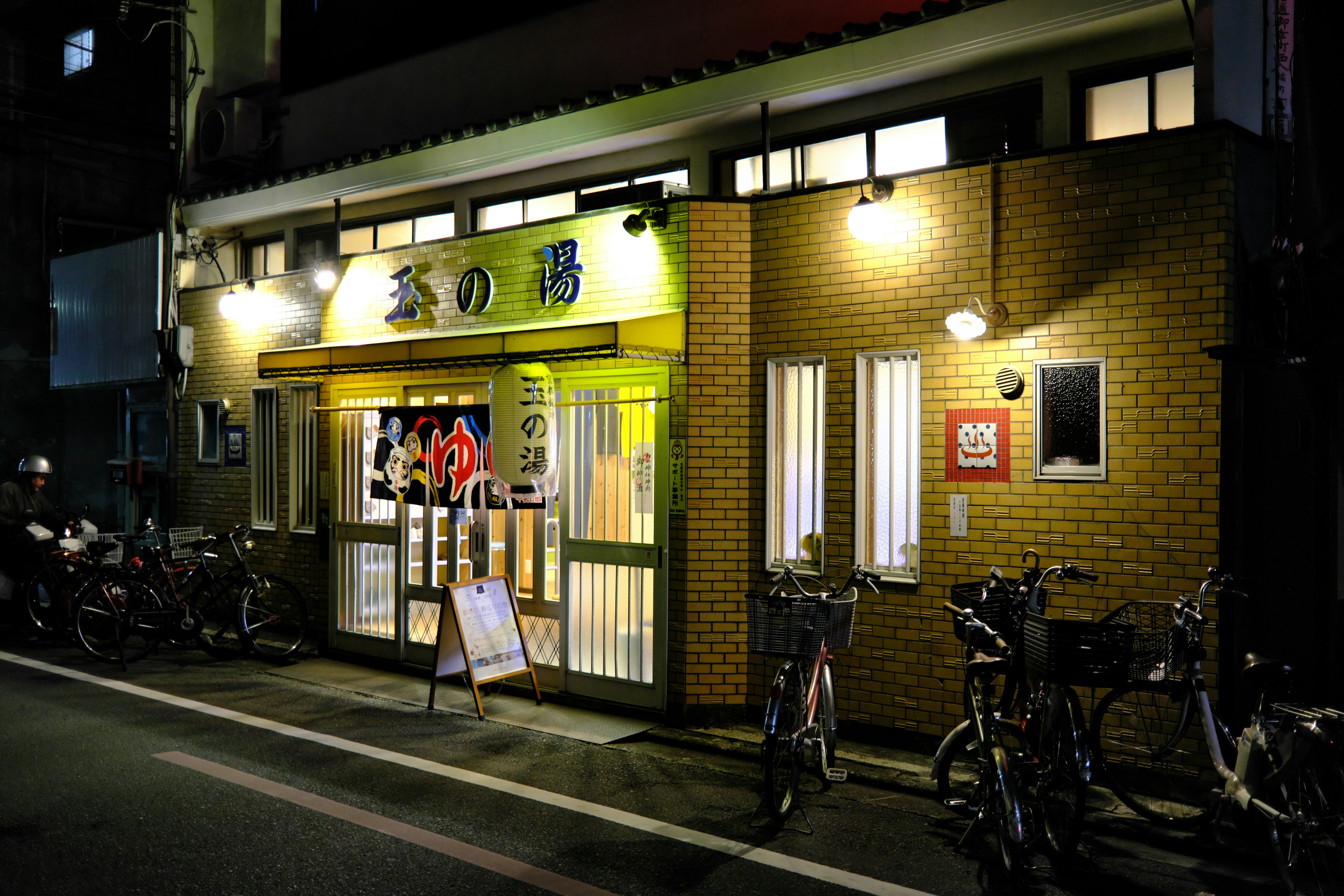 Exterior of an onsen at night featuring bright signage and parked bicycles