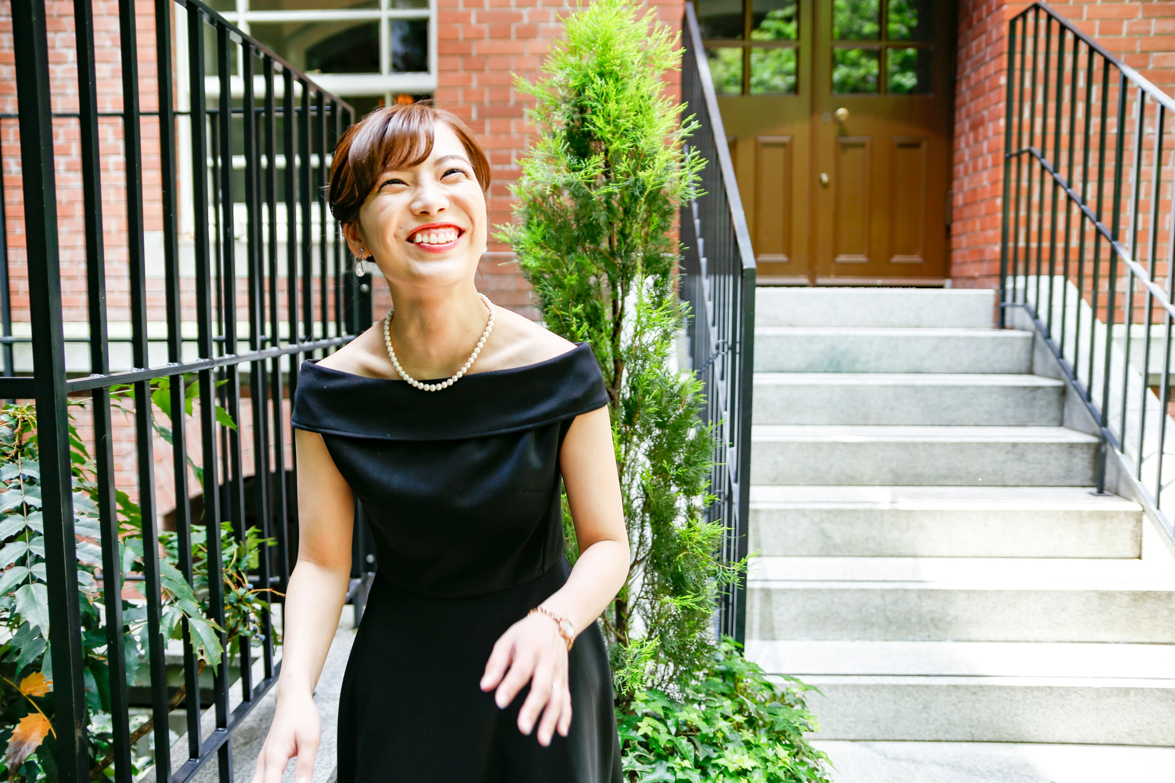 A woman in a black dress smiling standing near a brick building with greenery