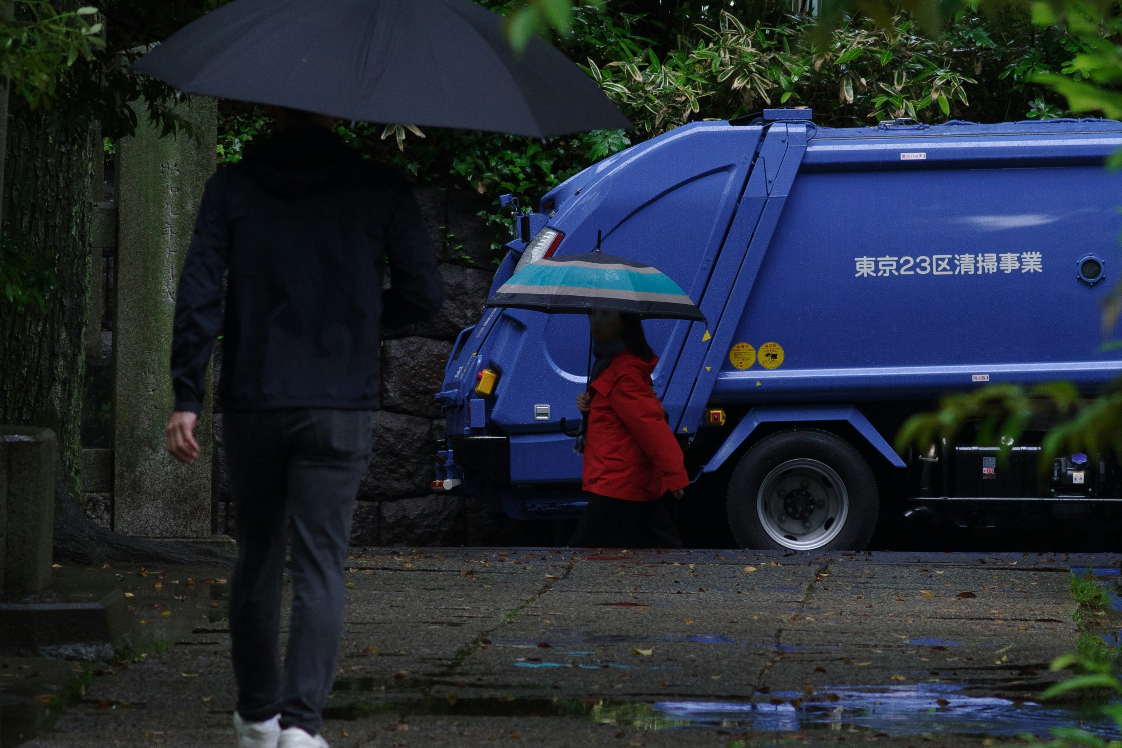 A person holding a black umbrella and a child with a red umbrella near a blue garbage truck in the rain