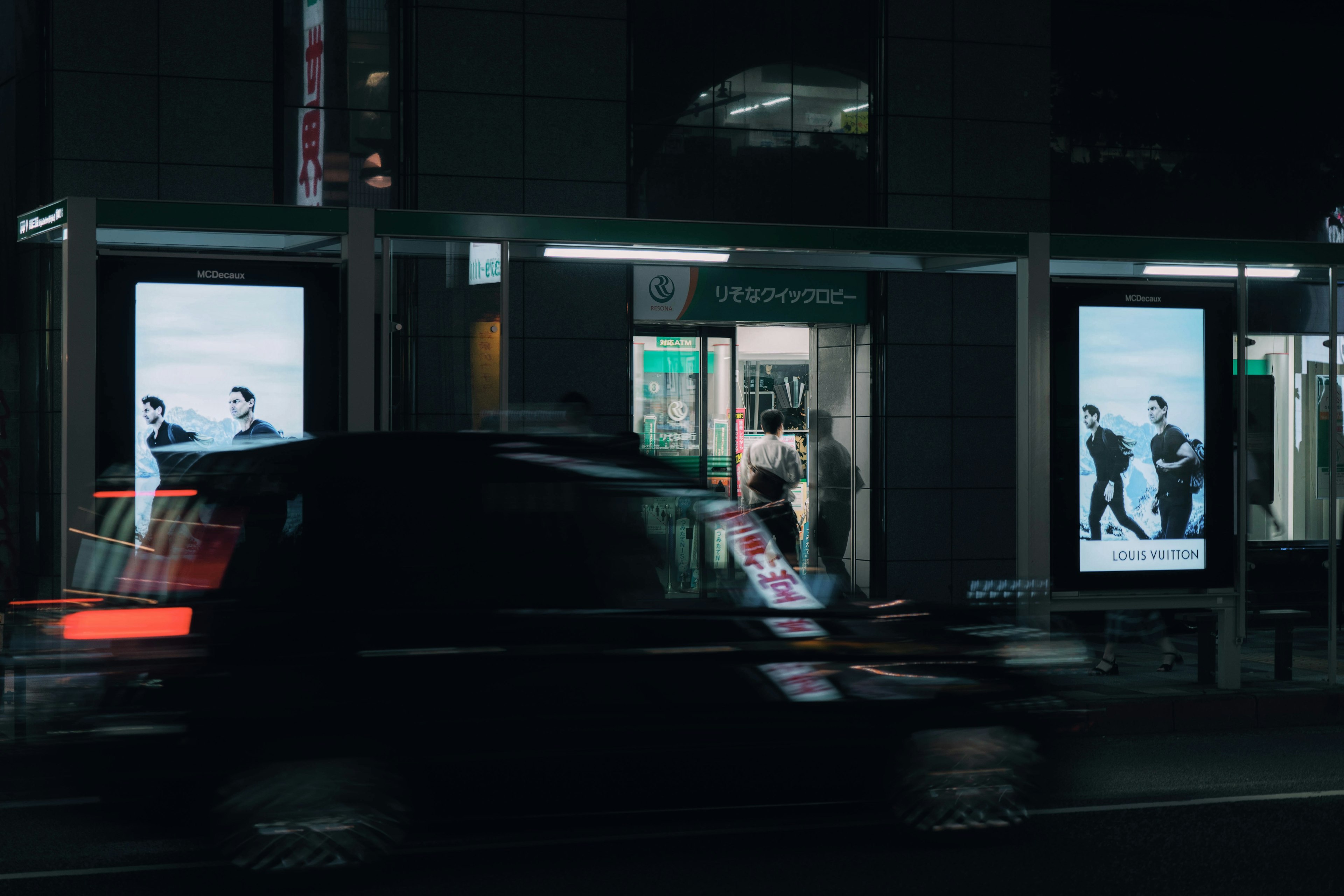 夜の街角に立つバス停と広告パネルが映るシーン