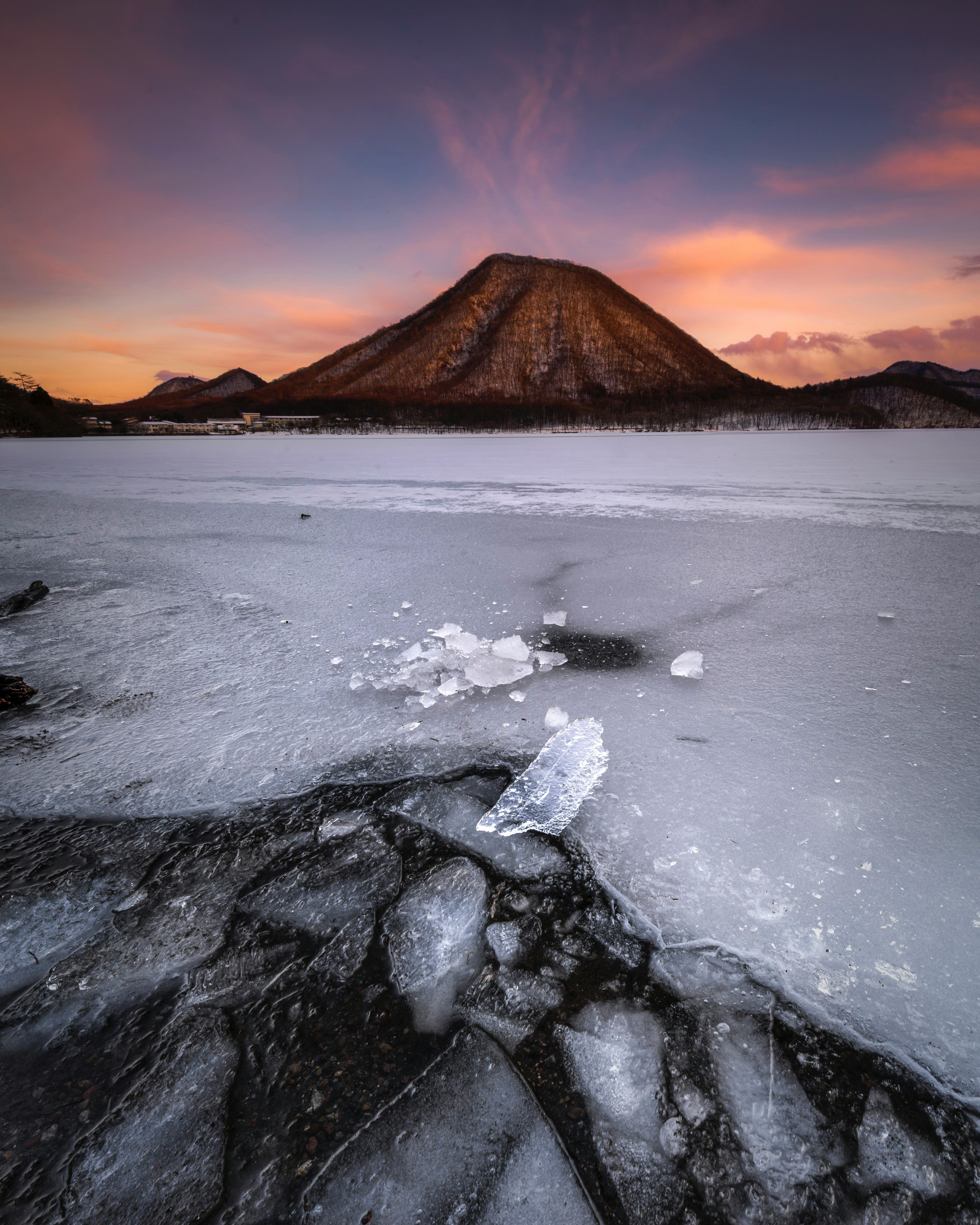 氷に覆われた湖の前景と背後にそびえる山のシルエットが美しい夕焼け