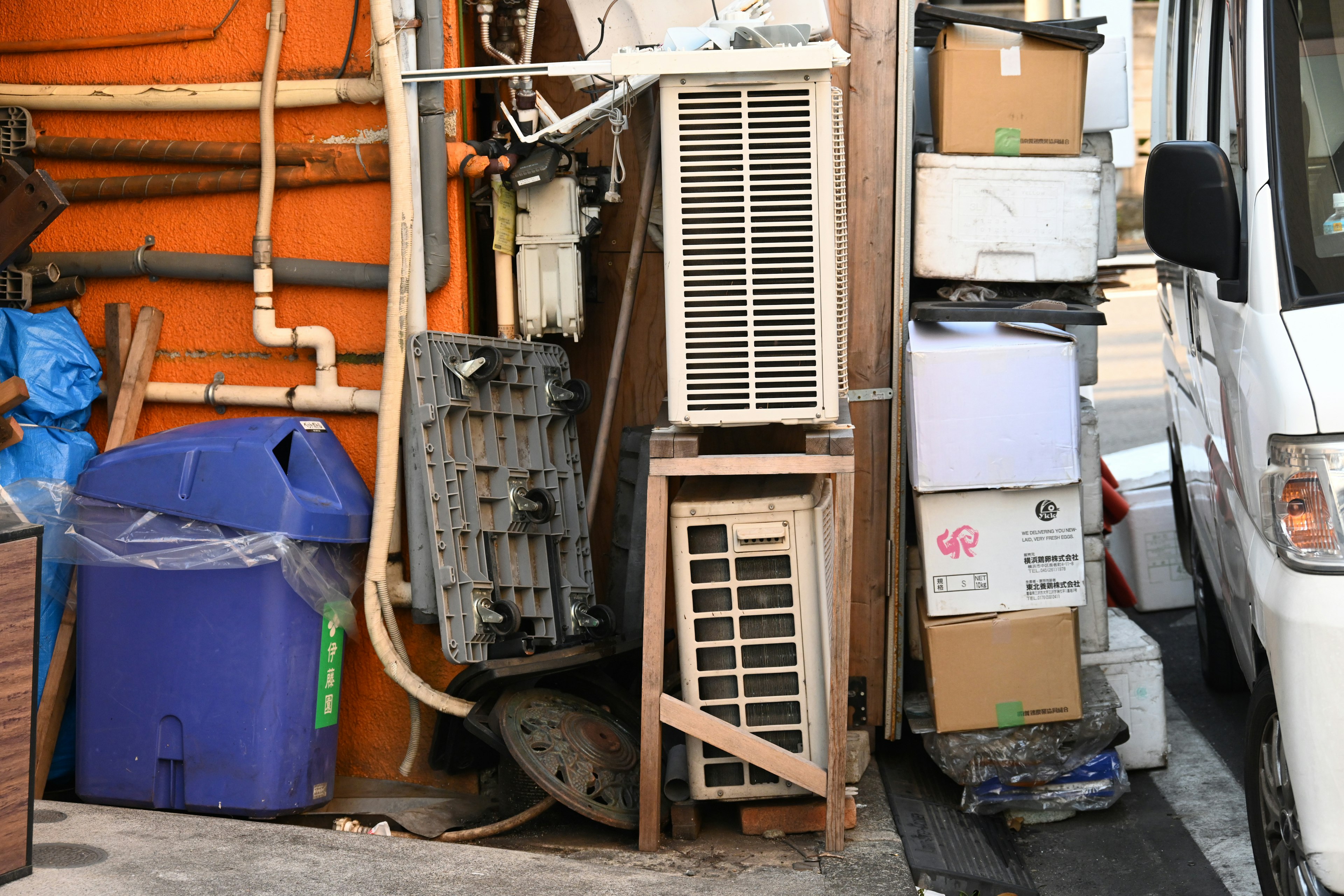 Narrow alleyway with an air conditioner and stacked cardboard boxes next to a trash bin