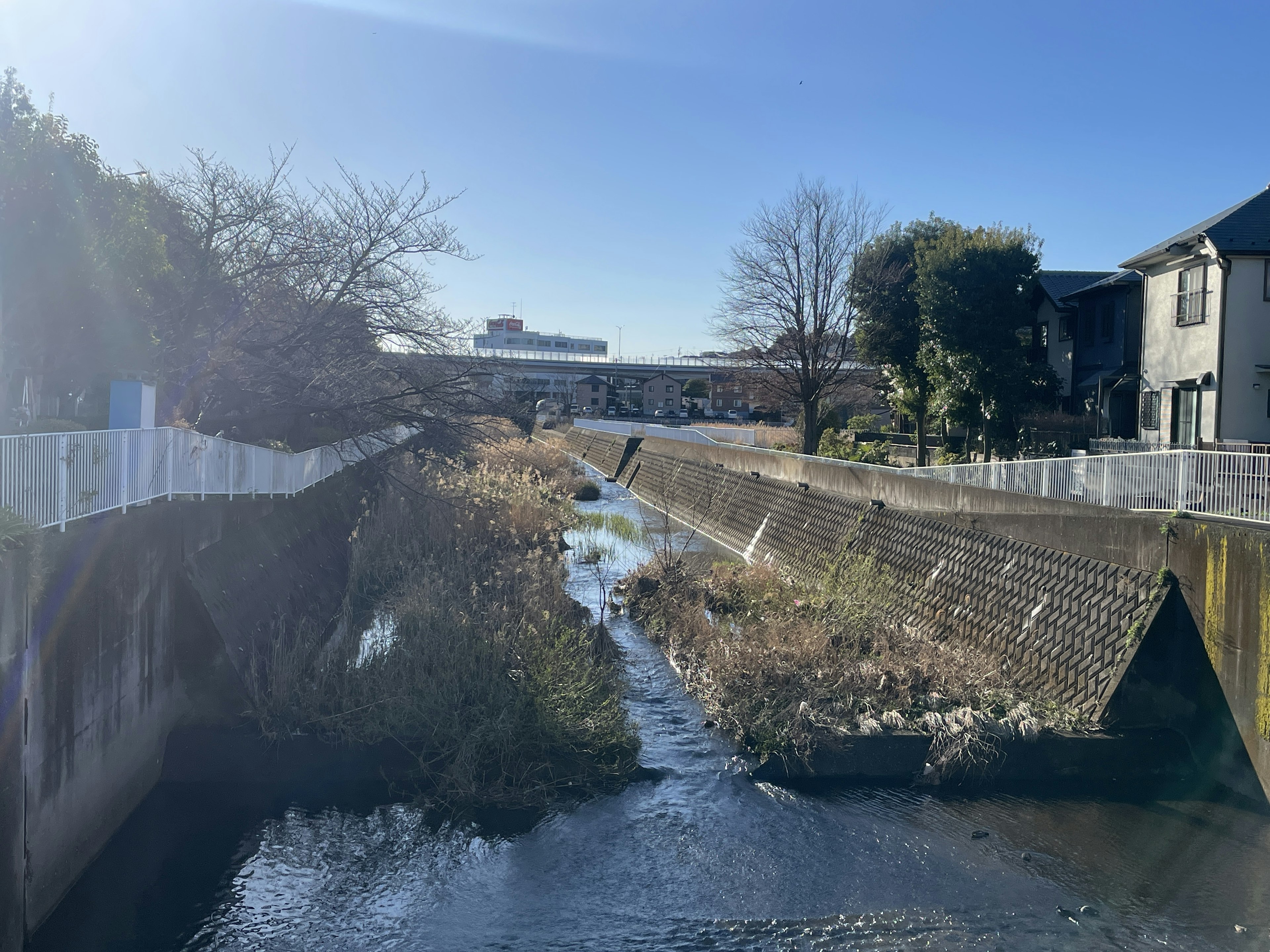 A small stream flowing under a blue sky with buildings and vegetation on both banks