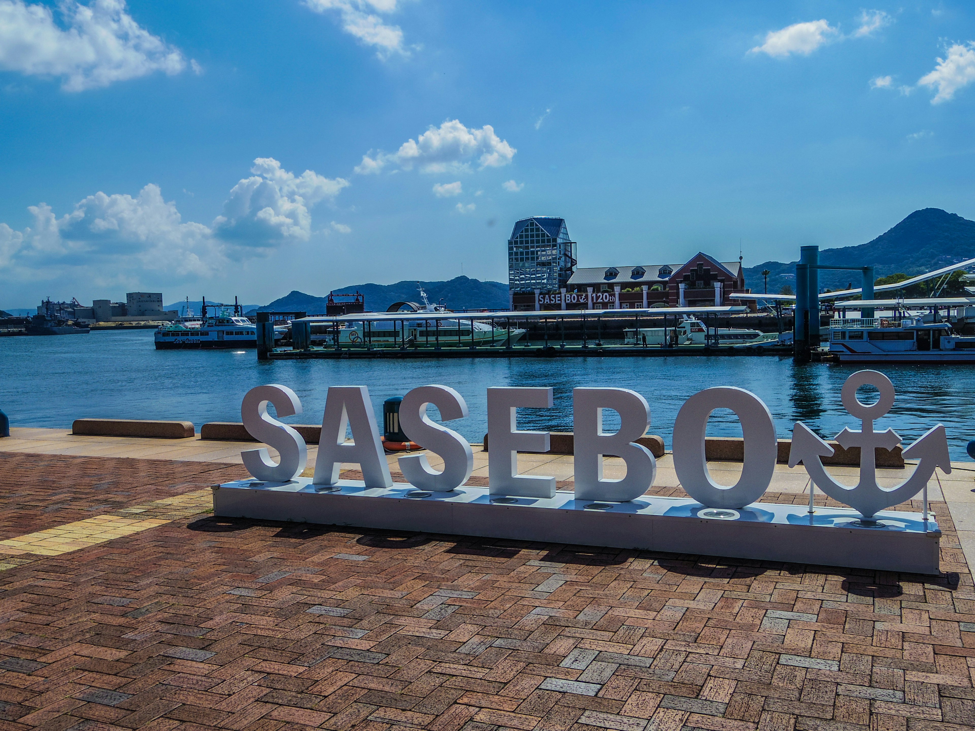 Tanda besar bertuliskan SASEBO di tepi laut dengan langit biru