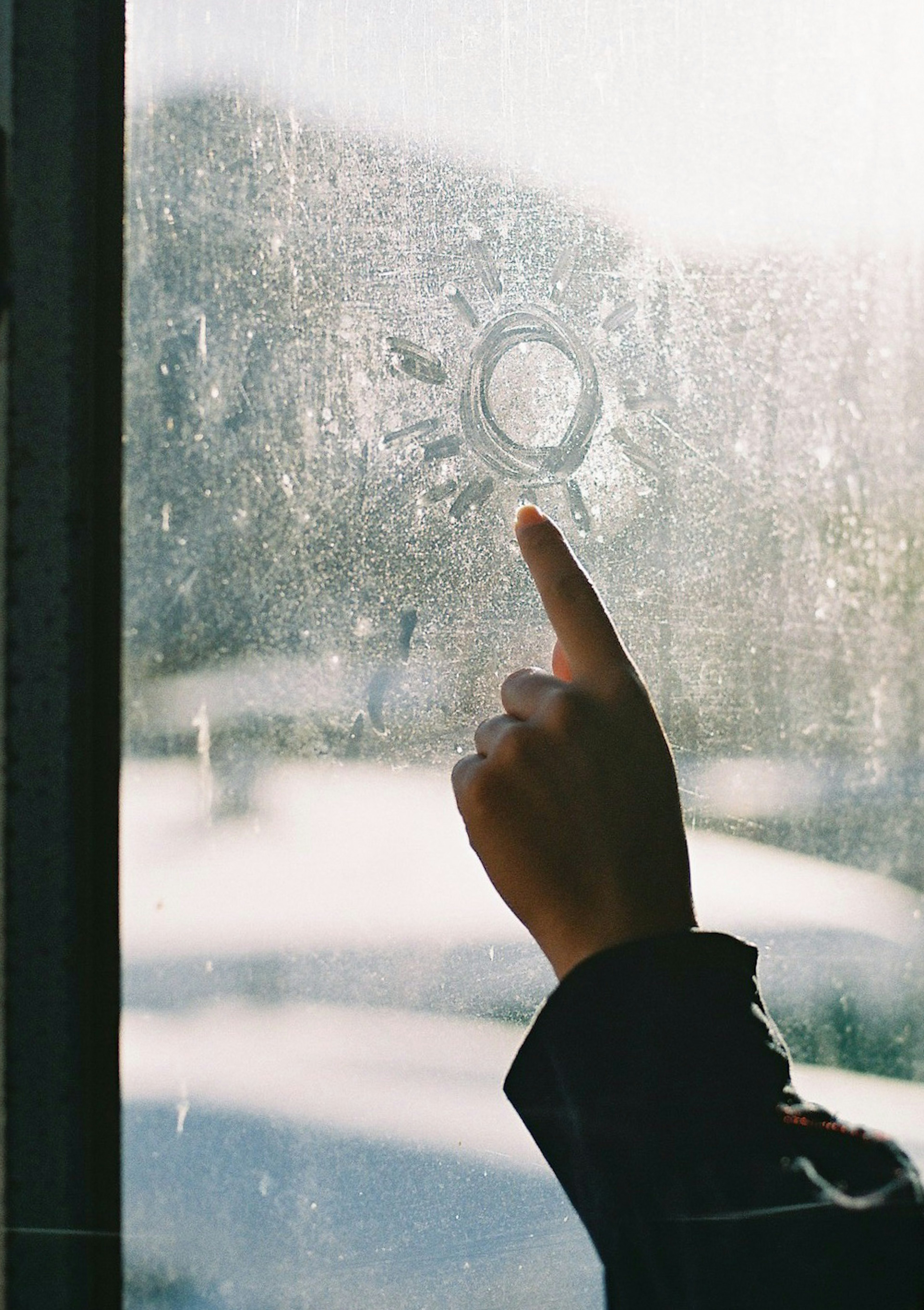 A hand tracing a circular pattern on a foggy window with sunlight in the background