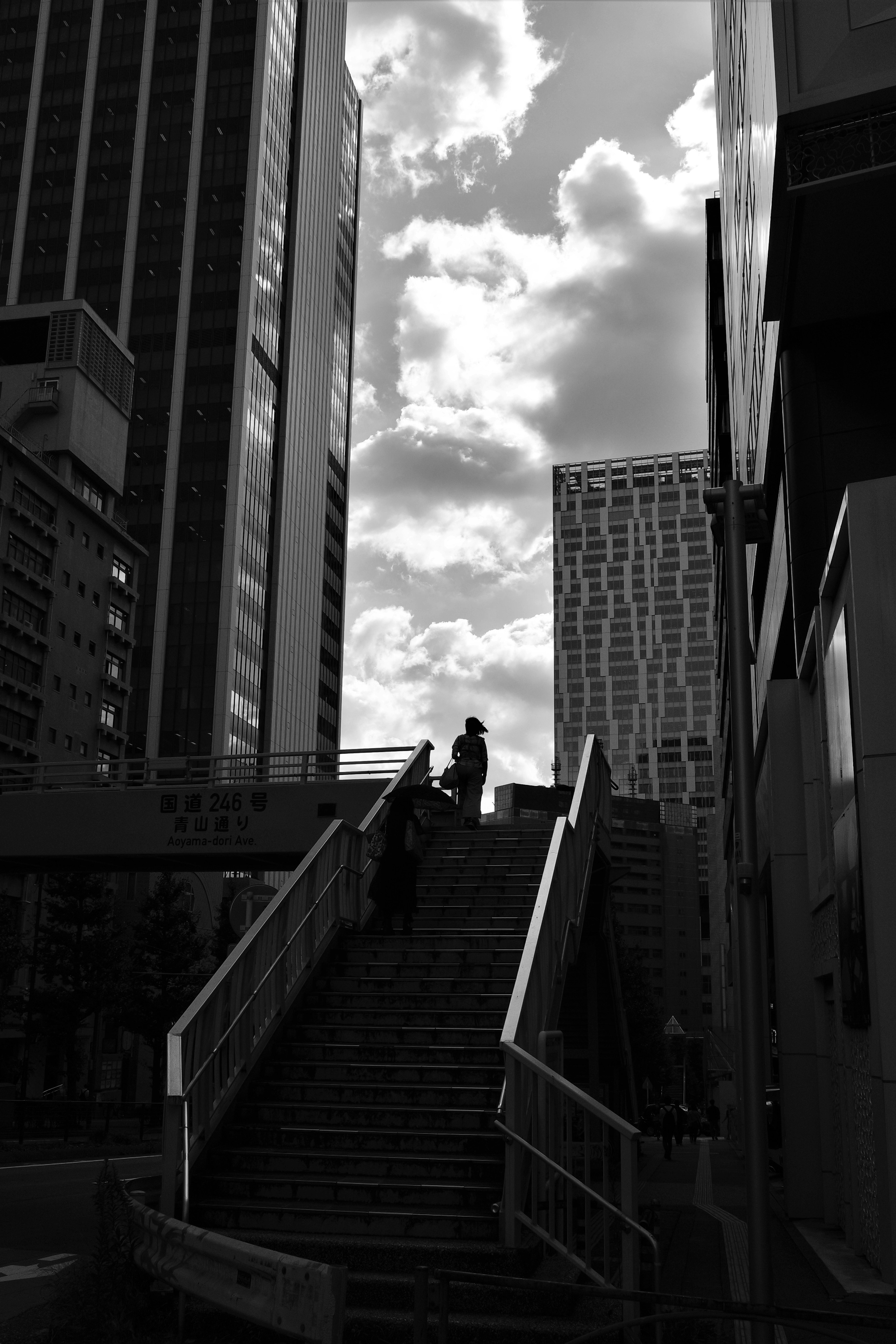 Monochrome staircase with people walking between buildings and a cloudy sky in the background