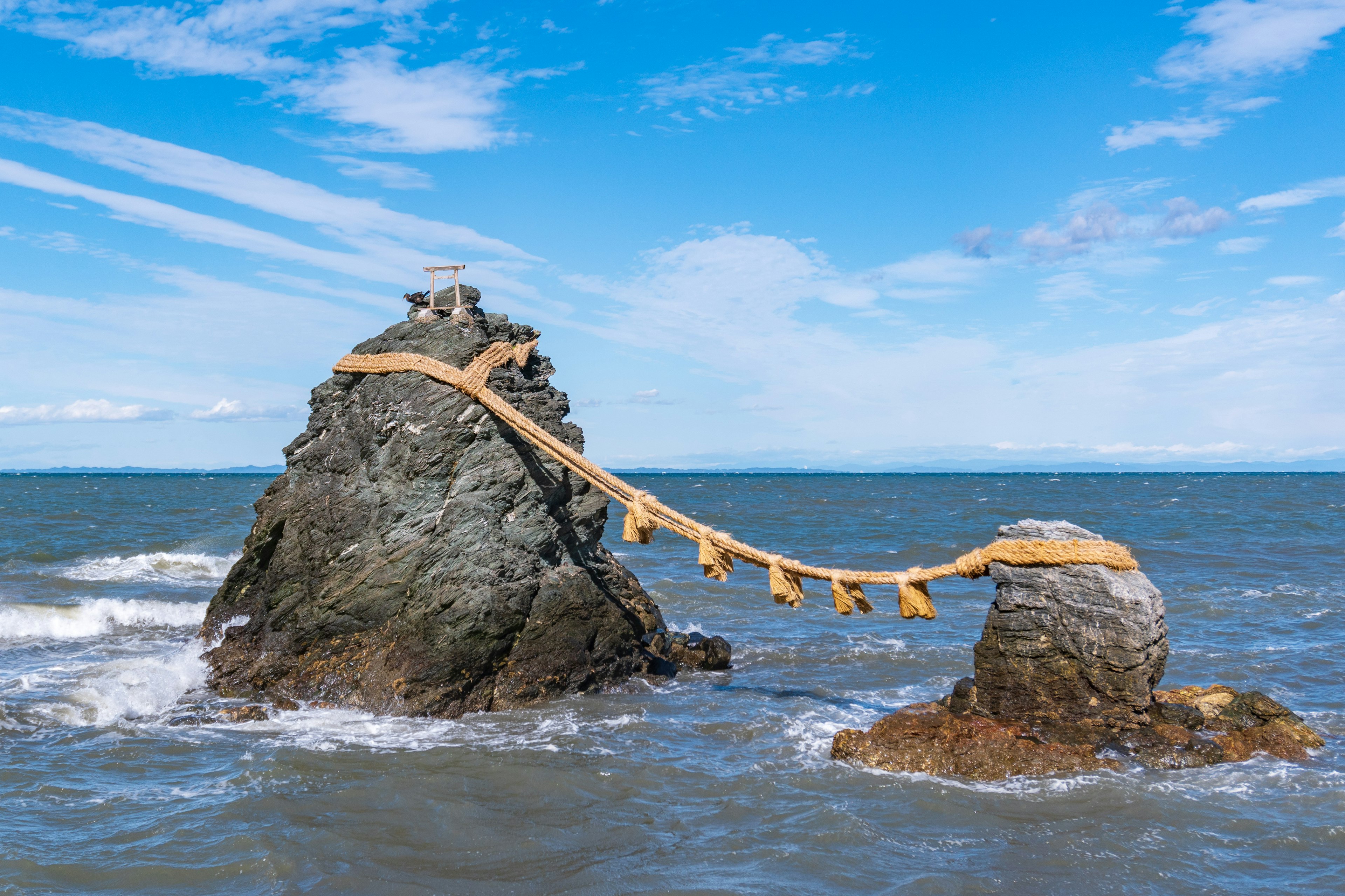 Dos rocas en el océano conectadas por un puente de madera bajo un cielo azul con nubes