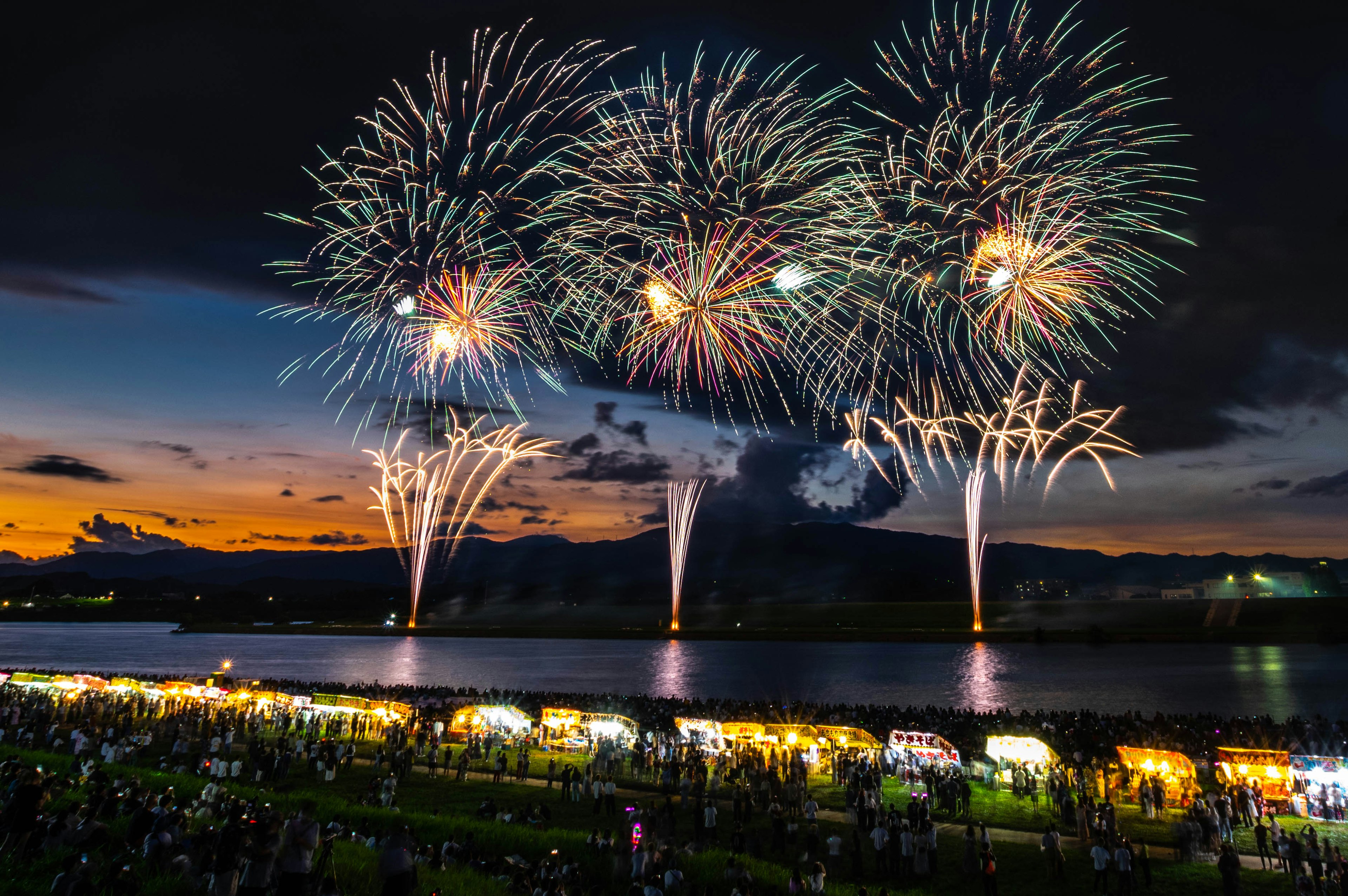 夜空に花火が打ち上げられ川のほとりに人々が集まる風景