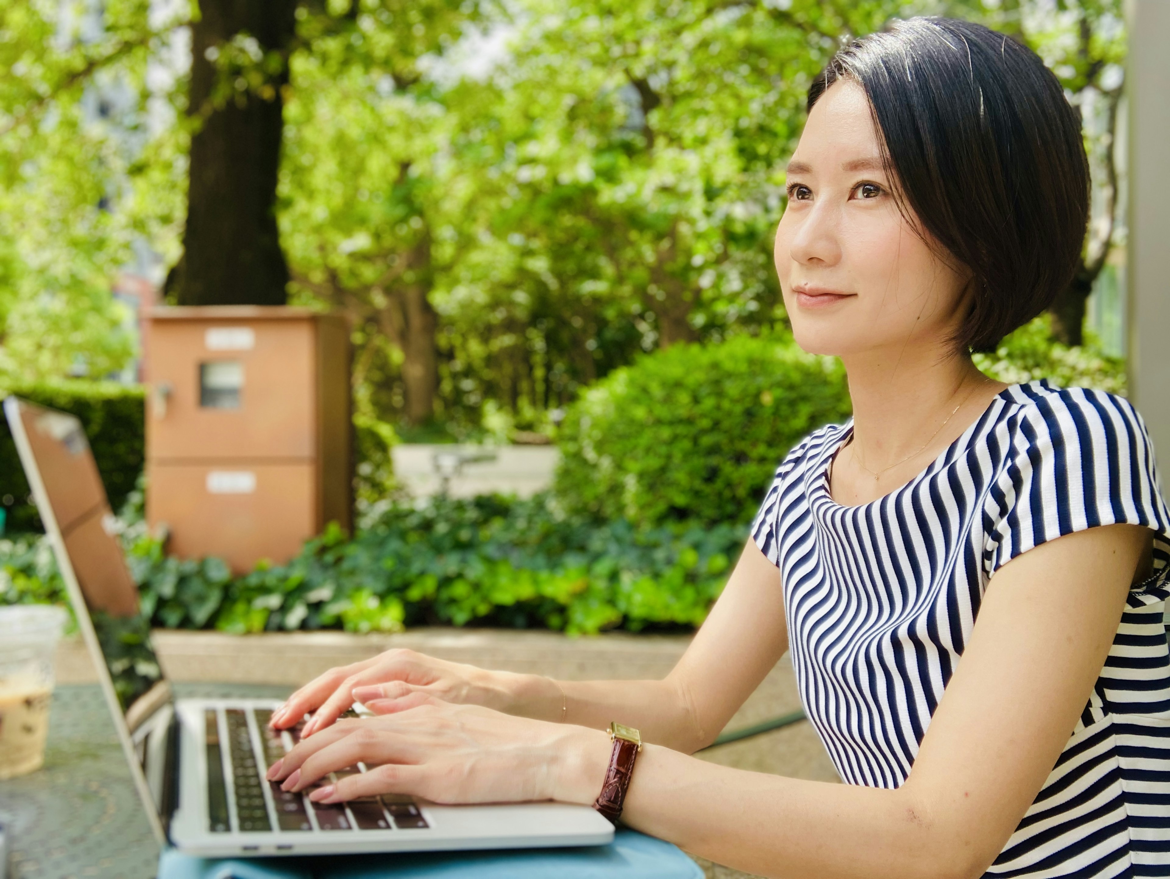 Frau, die in einem Park mit einem Laptop umgeben von Grün arbeitet und ein gestreiftes Shirt trägt