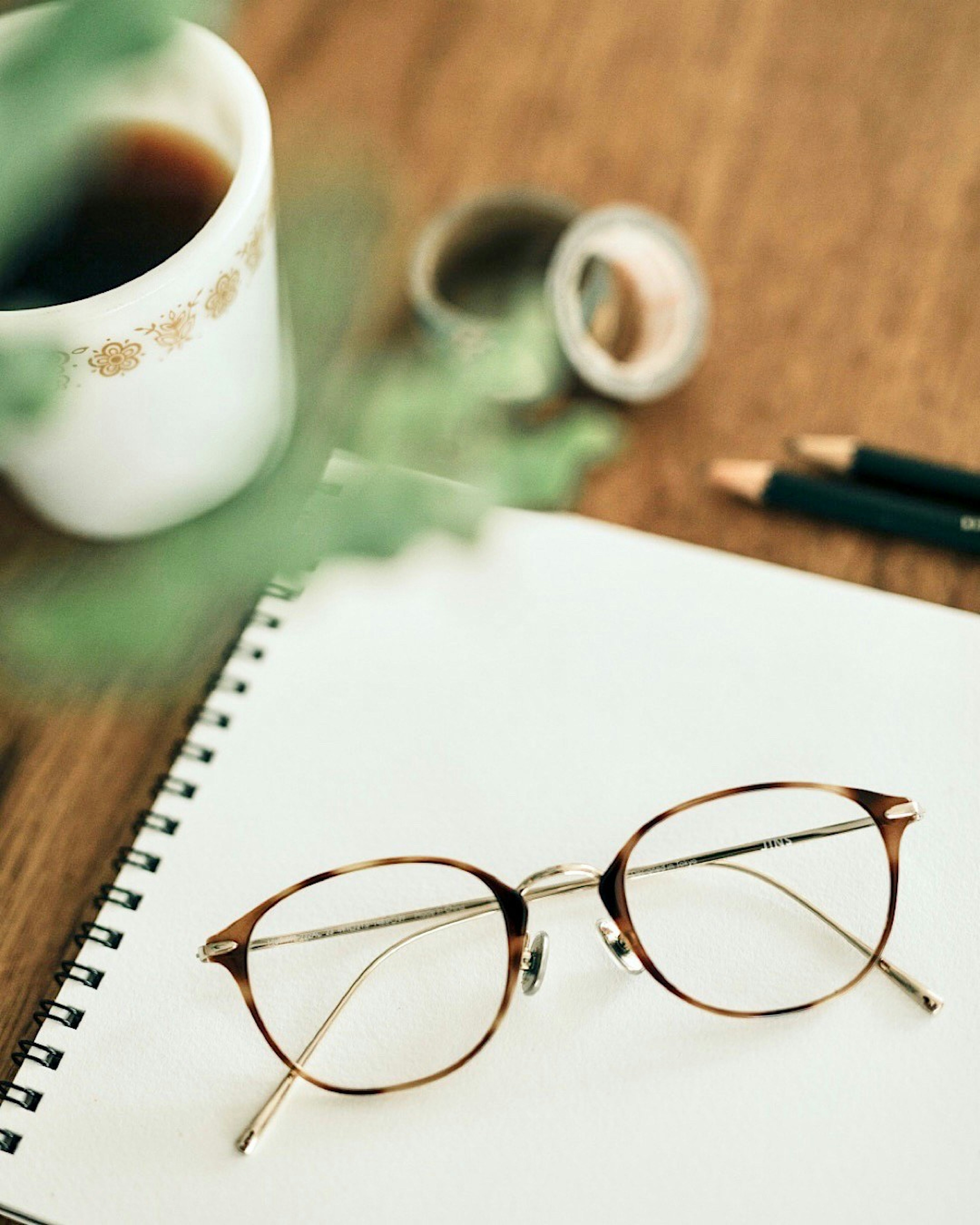 Brille und ein Notizbuch auf einem Holztisch mit einer Tasse Kaffee und einer Pflanze