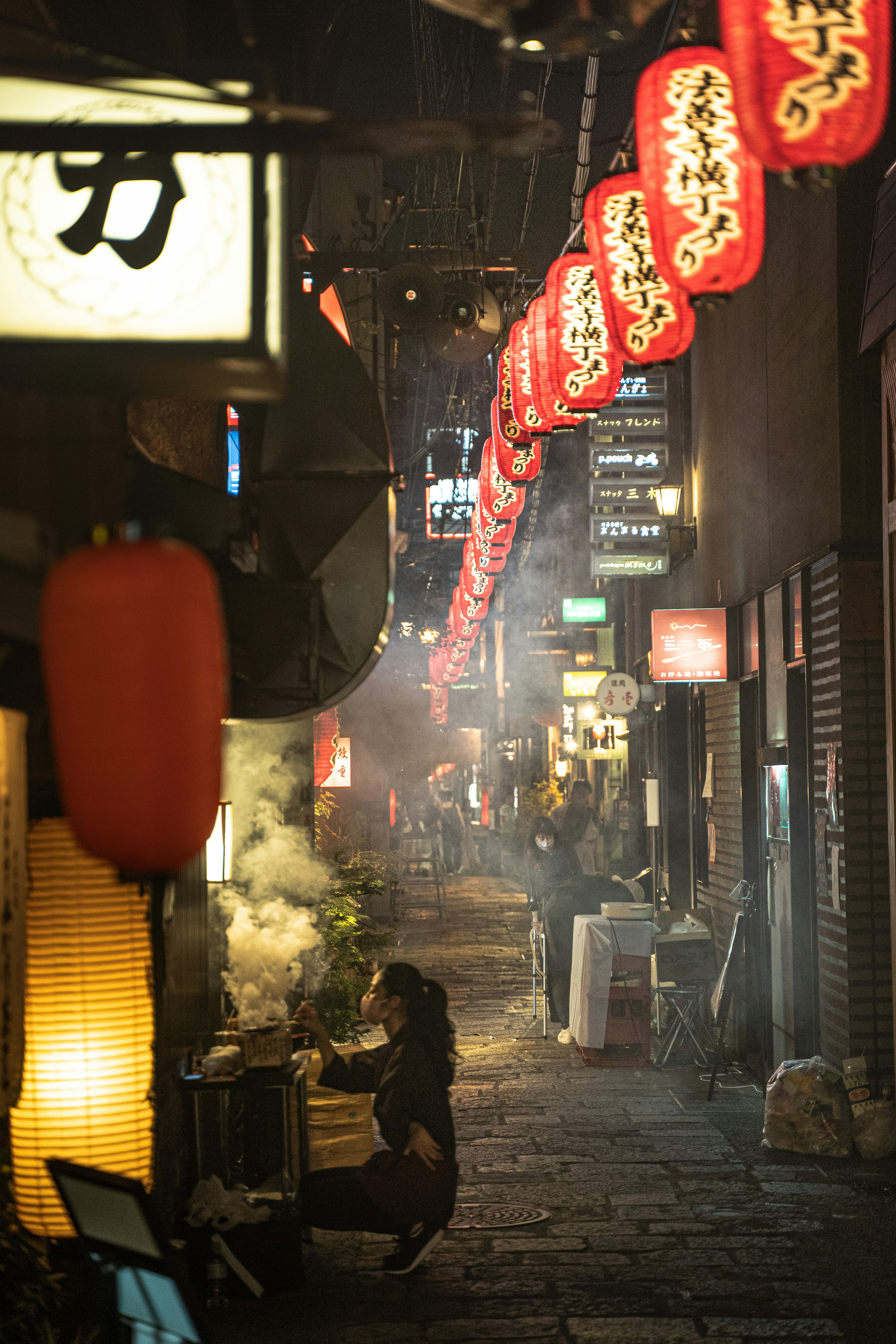 Narrow alley at night with hanging lanterns and a person cooking